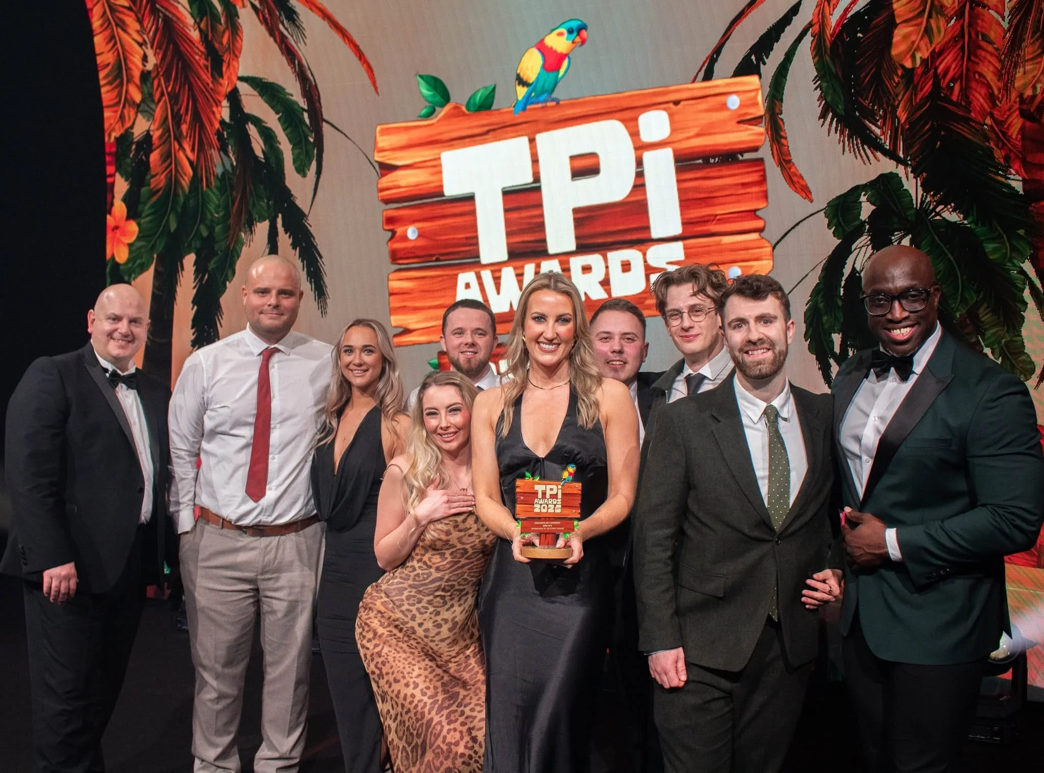 Group of nine people dressed in formal attire, standing in front of a tropical-themed backdrop with palm leaves and the text 'TPI Awards 2023.' One woman in the center holds a trophy.