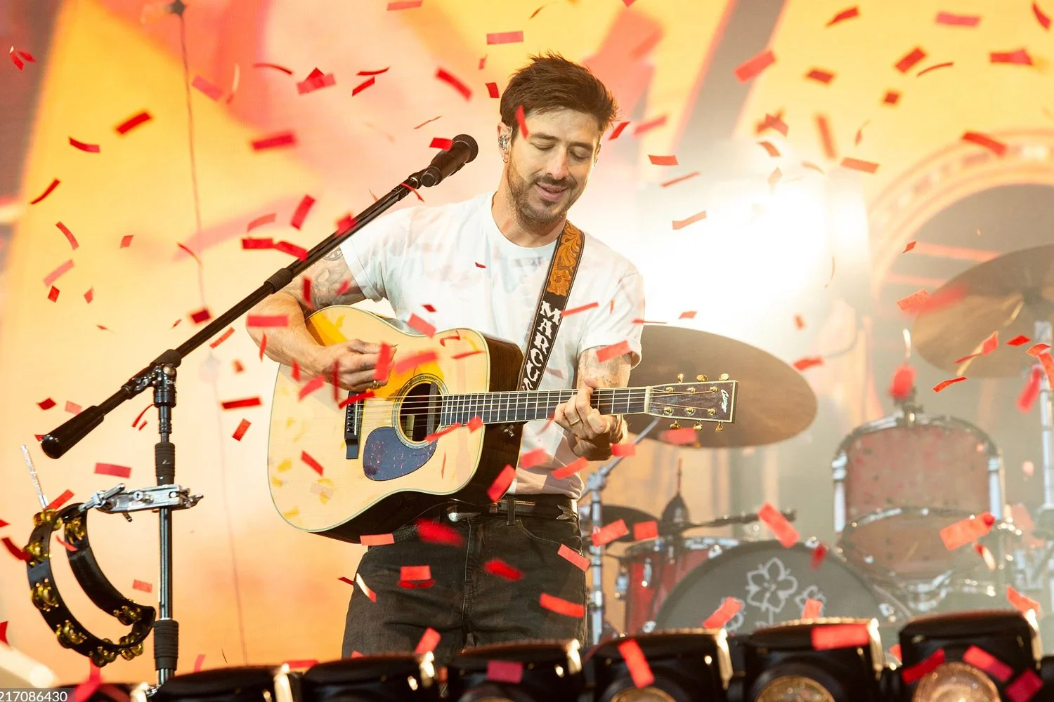 A man playing an acoustic guitar on stage, surrounded by red confetti, with a drum set behind him and bright stage lights.
