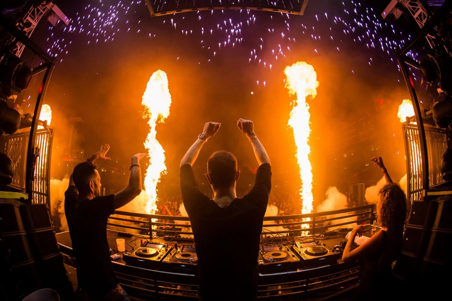 A DJ performing at a concert with fire and pyrotechnics on stage, audience members dancing and raising their hands.