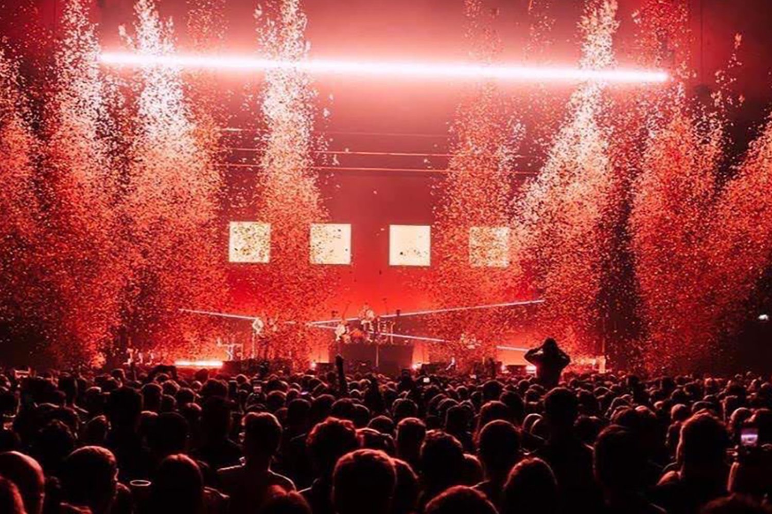 A concert with a large crowd, bright red stage lighting, and pyrotechnics or fireworks during the performance.