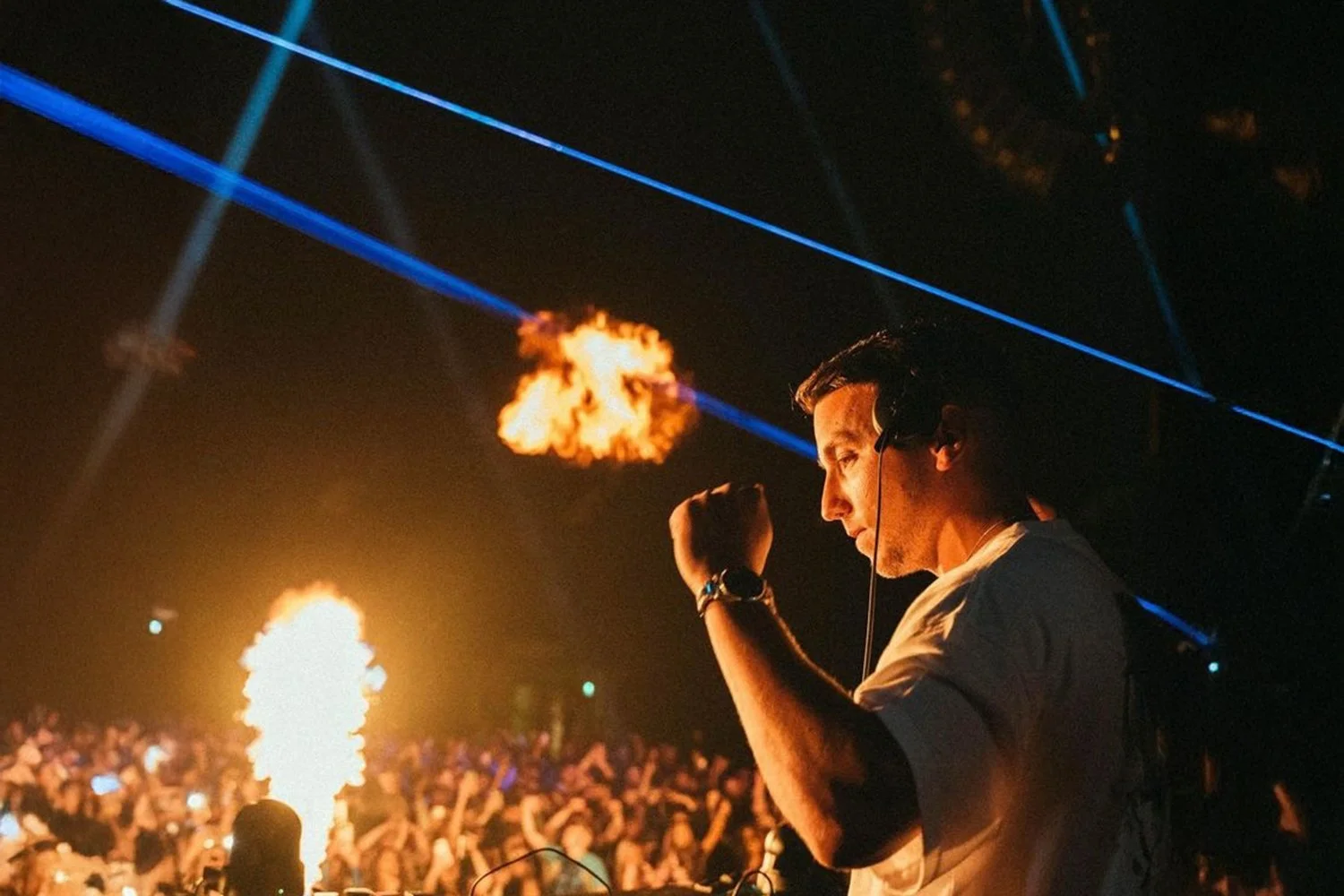 A male DJ wearing headphones performs at a nightclub with vibrant blue laser lights, flames shooting in the background, and a large crowd dancing.