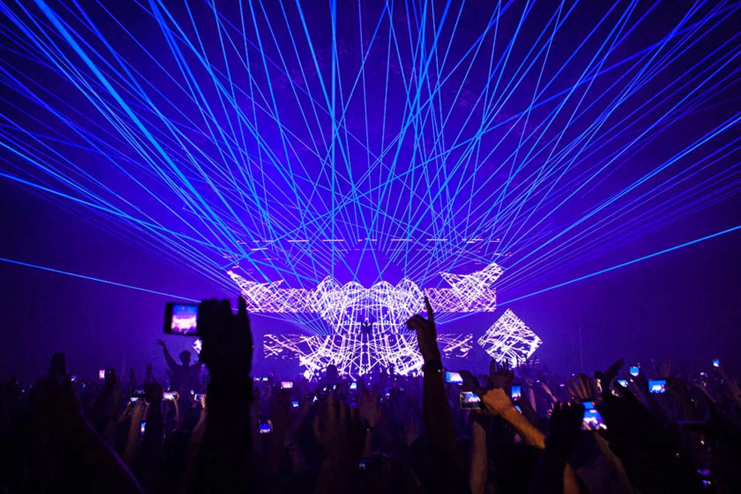A large crowd at a concert or music event with many people taking photos with smartphones. The stage features a vibrant display of blue and purple laser lights and digital visuals creating a dynamic, energetic atmosphere.