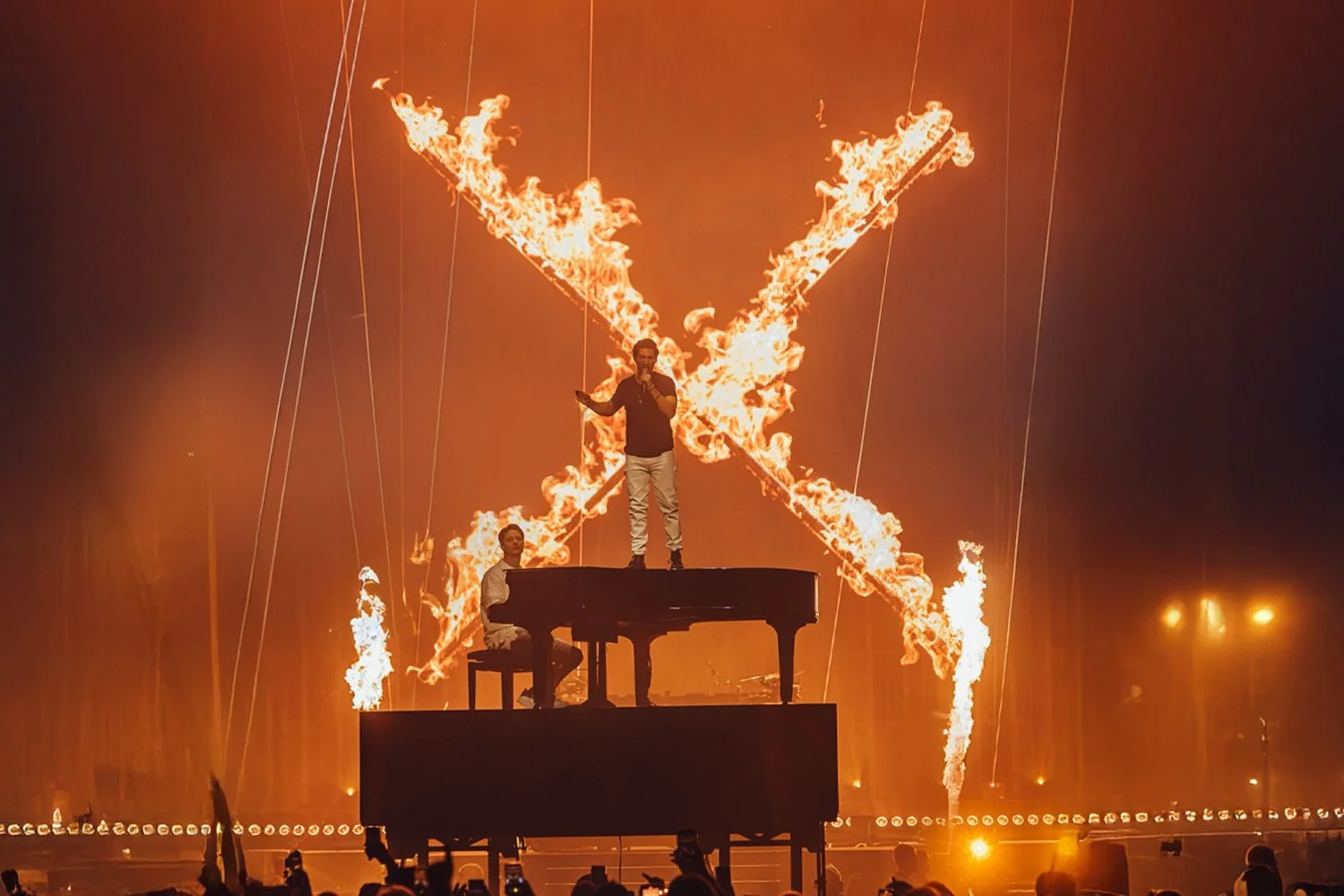 Performer standing on a raised platform behind a piano, with another performer sitting at the piano, during a fire show with fire effects in the shape of a butterfly in the background.