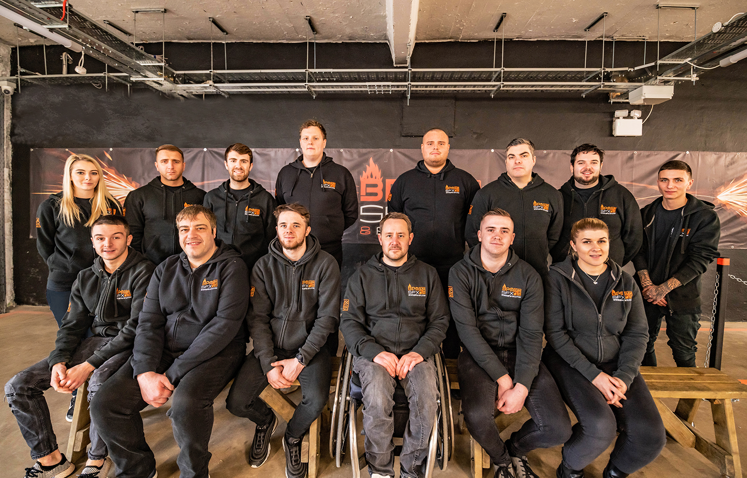 Group portrait of 13 young adults, men and women, wearing black hoodies with a logo, posing in an indoor setting with a black wall and a banner in the background, some sitting on benches and others standing.