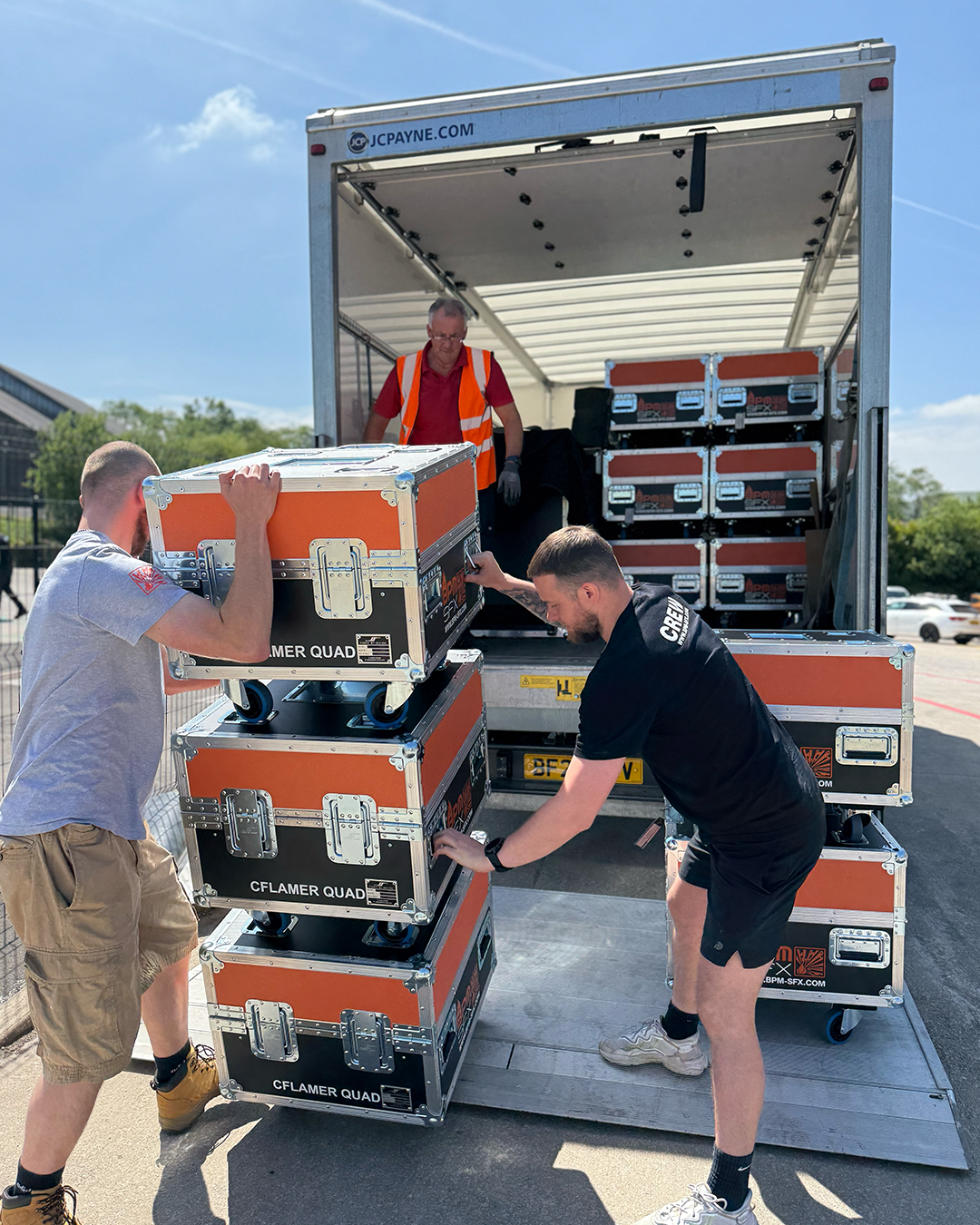 Three men unload orange and black equipment cases from a moving truck on a sunny day. One man is inside the truck, and two are outside moving the cases.