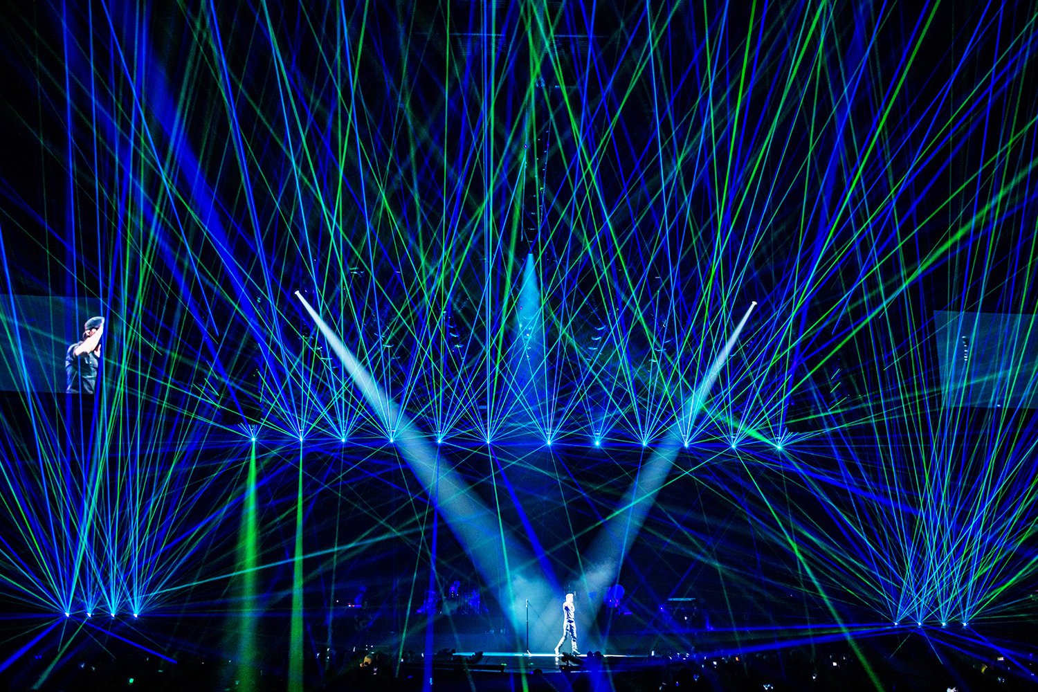 Concert scene with performer on stage, surrounded by blue and green laser lights forming an intricate pattern.