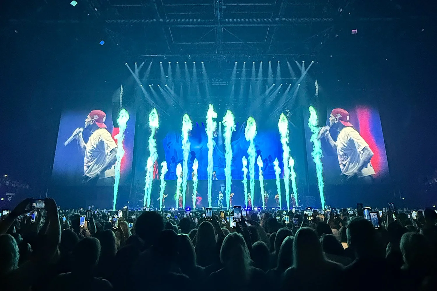 A concert scene with a performer on stage wearing a red cap, yellow shirt, and holding a microphone, shown on large screens on either side. The stage features colorful fireworks or pyrotechnics, with numerous audience members taking photos with their phones.