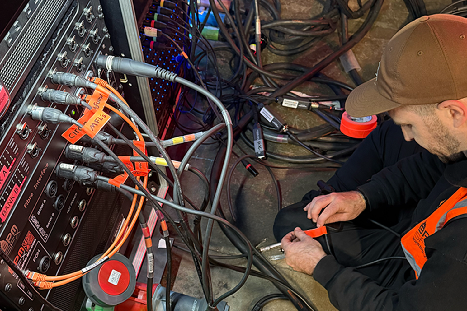 A man working on electronic equipment with numerous cables and wires, wearing a brown cap and an orange safety vest, sitting on the floor.