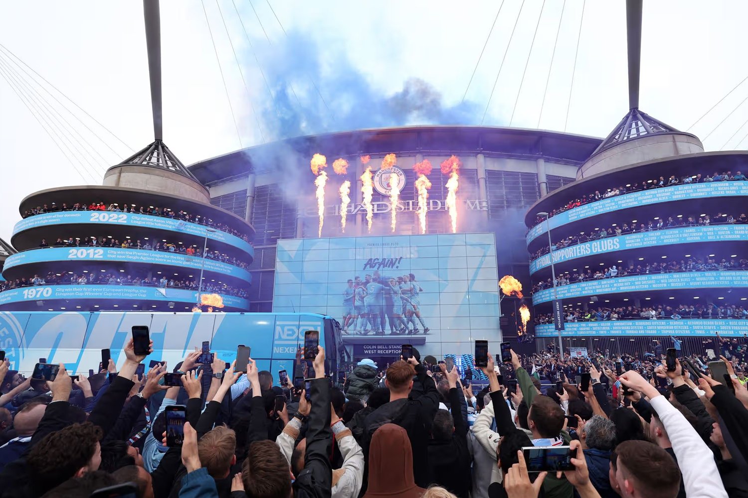 Crowd gathered outside a stadium with fire effects, fireworks, and a large screen displaying a team of sports players celebrating, during a sporting event.