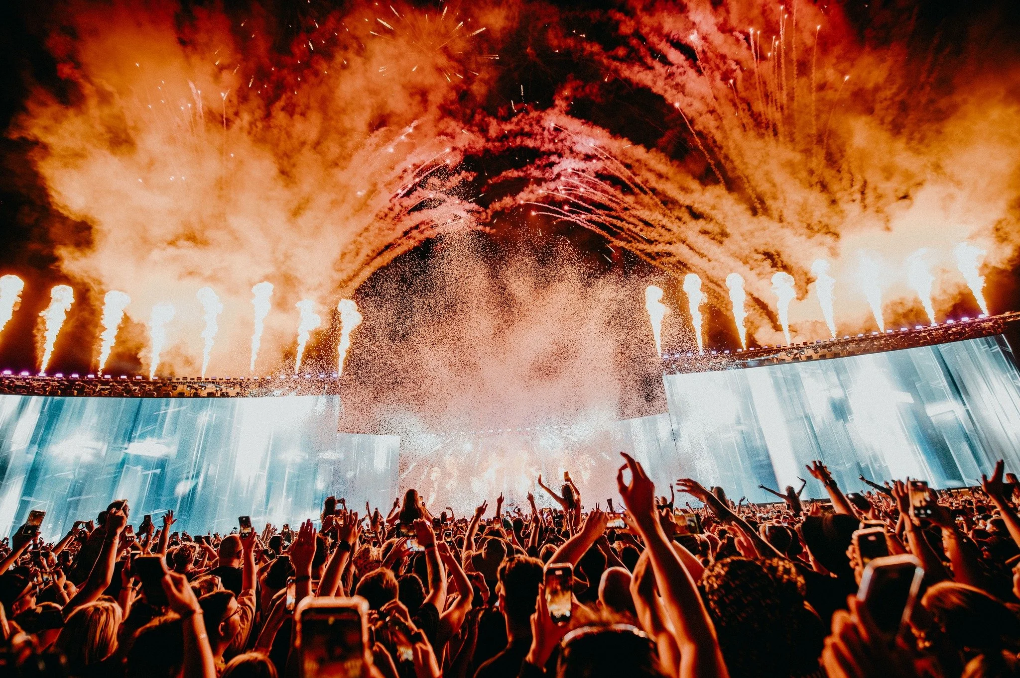 Crowd of people at a concert with fireworks and smoke in the background, raising their hands and taking photos.