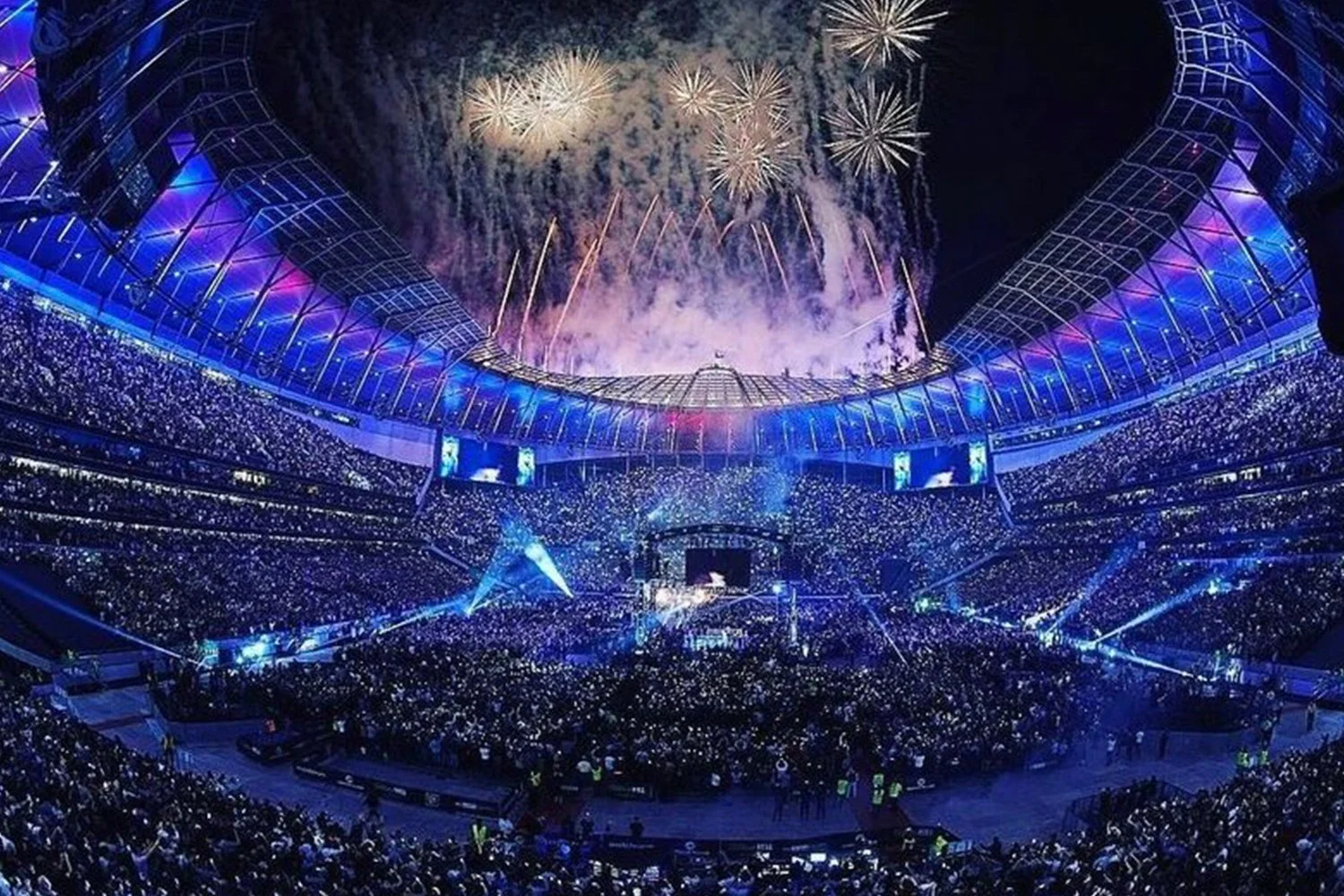 Large stadium filled with spectators watching fireworks above the stadium at night, illuminated with blue lights.