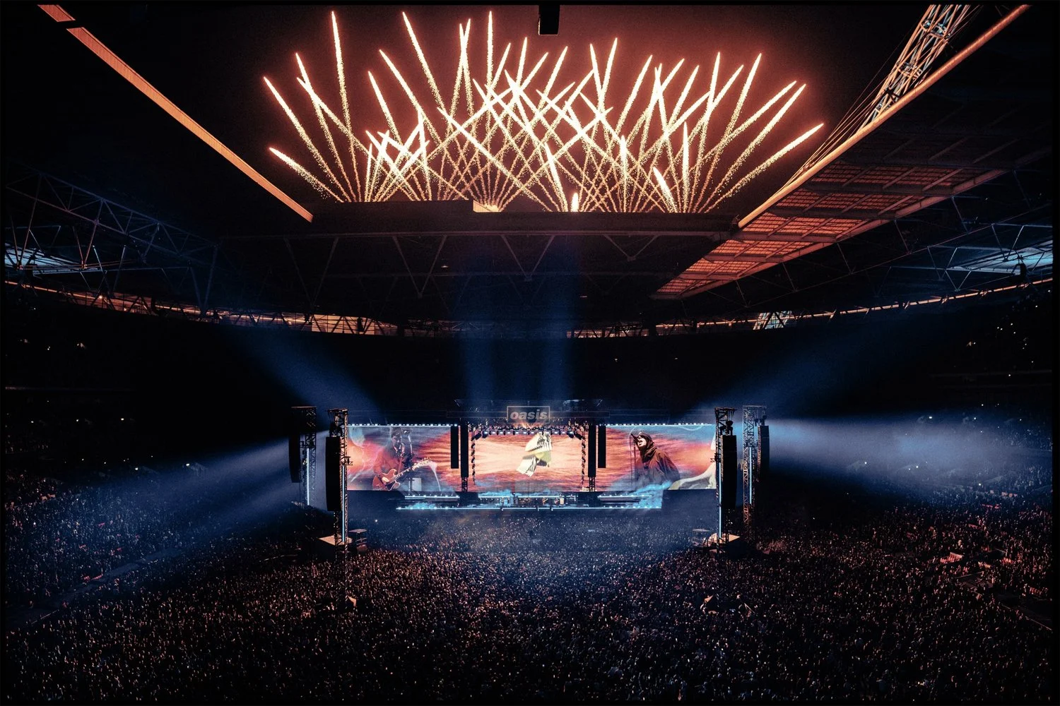 A large concert stadium filled with a crowd watching a stage performance, with multiple screens displaying performers, and fireworks lighting up the night sky above.