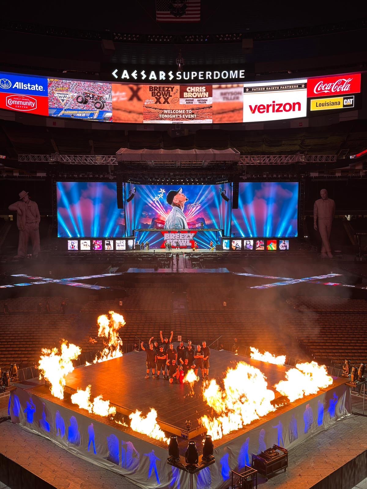 View of Caesars Superdome interior during Breezy XX event, with large stage, fire effects, giant screens, and personnel preparing on stage.
