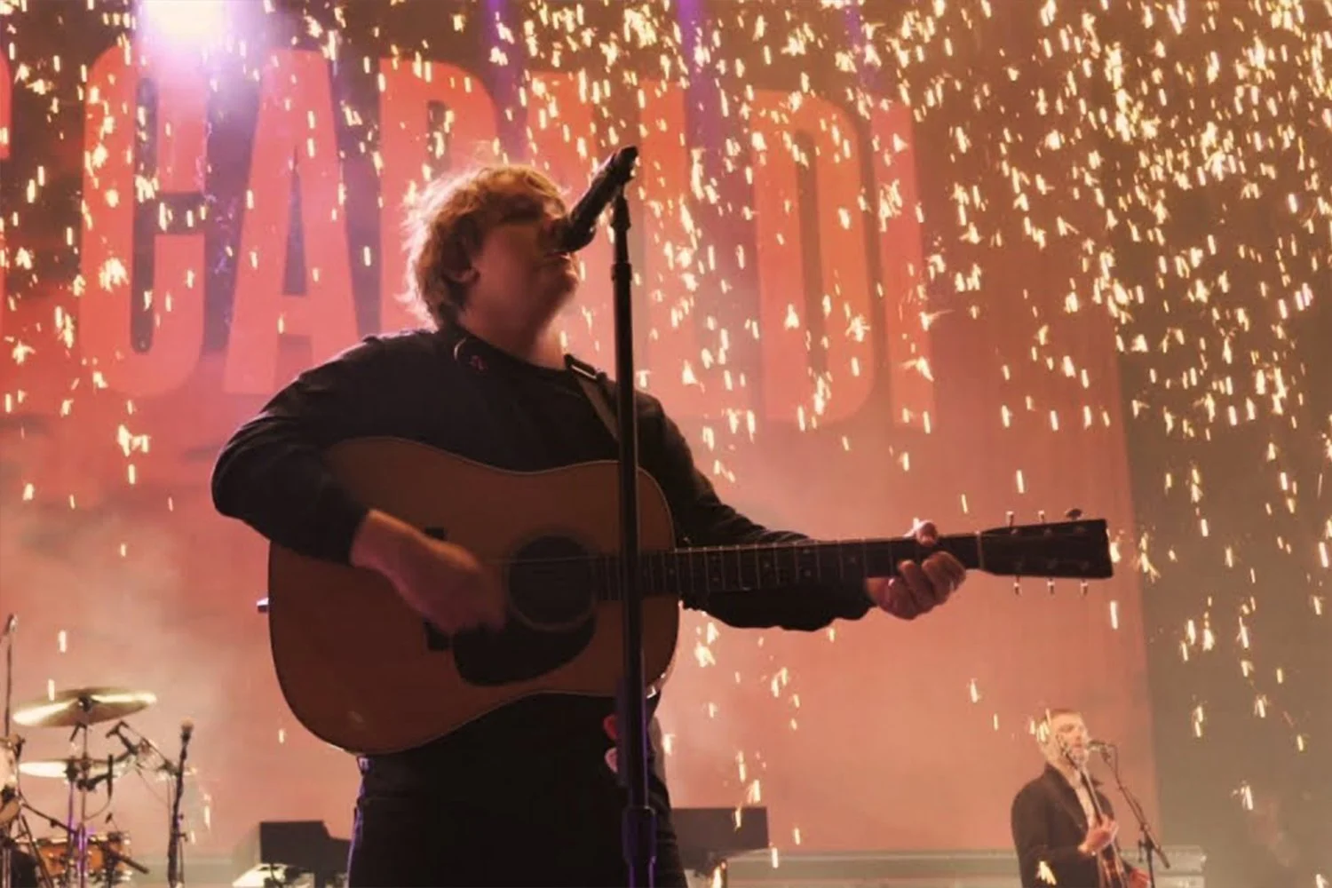 Person playing acoustic guitar and singing into microphone during a concert with fireworks display in the background.