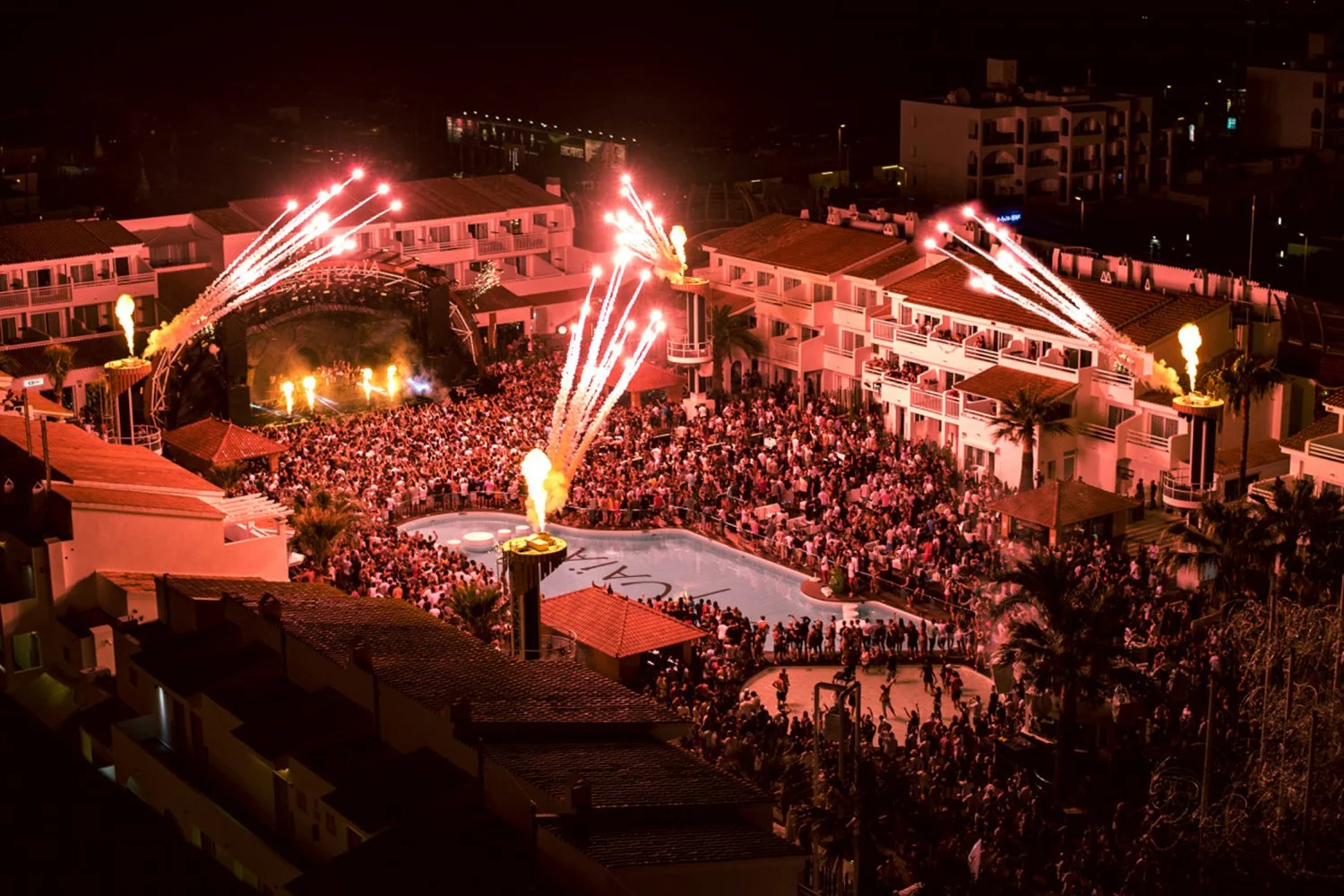 Nighttime scene of a crowded outdoor party at a resort with fireworks, fire breathers, palm trees, and a swimming pool, surrounded by buildings.