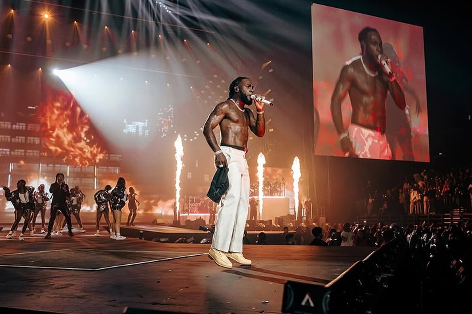 A shirtless male performer with long hair, wearing white pants and beige sneakers, stands on stage holding a microphone, with pyrotechnics and flames in the background, and a large screen displaying his image.