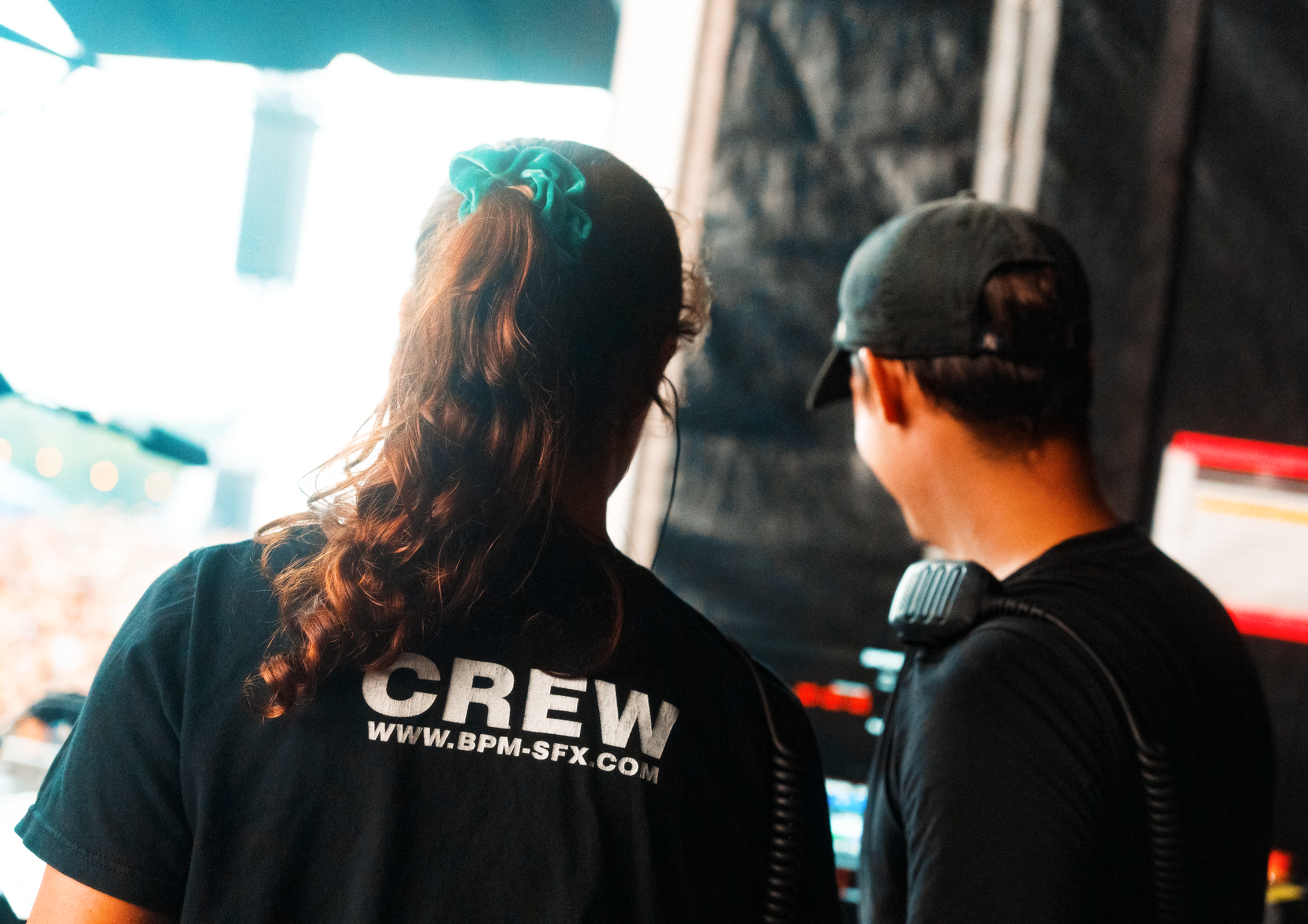 A woman and a man standing close together, viewed from behind. The woman has long, curly hair tied with a teal scrunchie and is wearing a black t-shirt that reads 'CREW' and includes a website URL. The man is wearing a black cap and a black shirt with a visible arm tattoo. They are indoors, possibly in a store or cafe, with bright light coming from a window or entrance in the background.