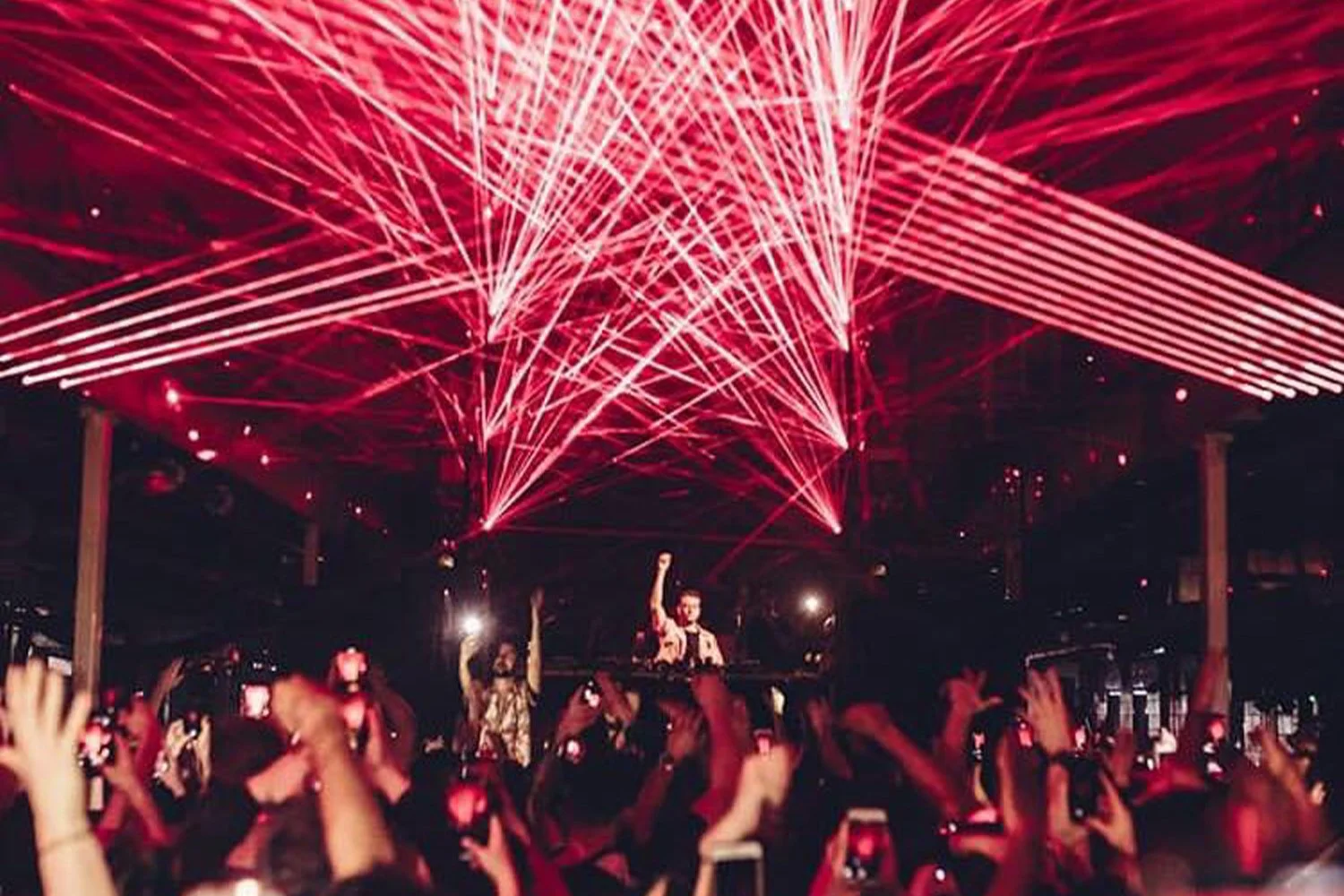 A concert scene with a DJ and a crowd of people raising their hands, illuminated by red laser lights and fireworks.