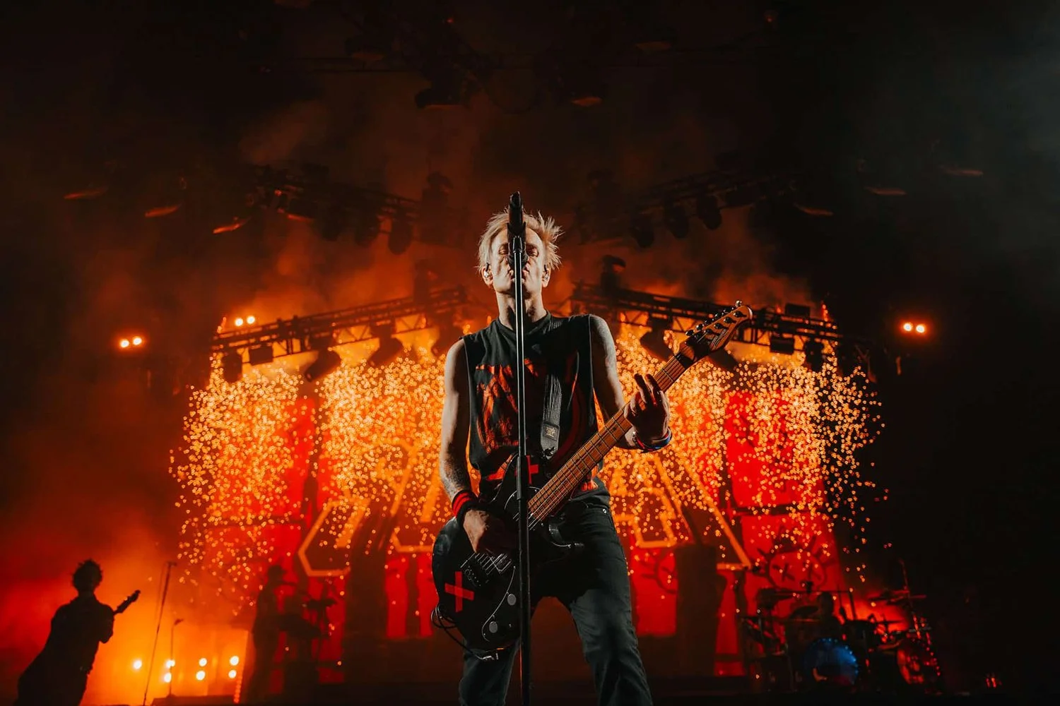 Musician performing on stage with guitar, illuminated by red and orange lights, with a background of fire and pyrotechnics.