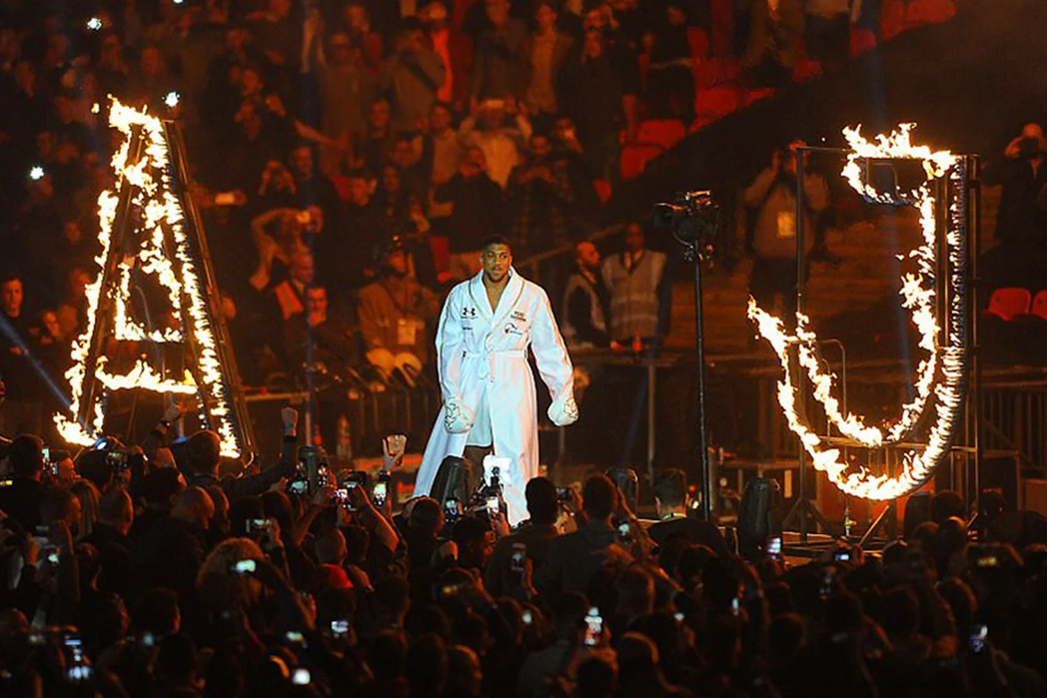 A male athlete in a white judo uniform standing on stage with flames surrounding large letter U, with an audience taking photos and videos in the background.
