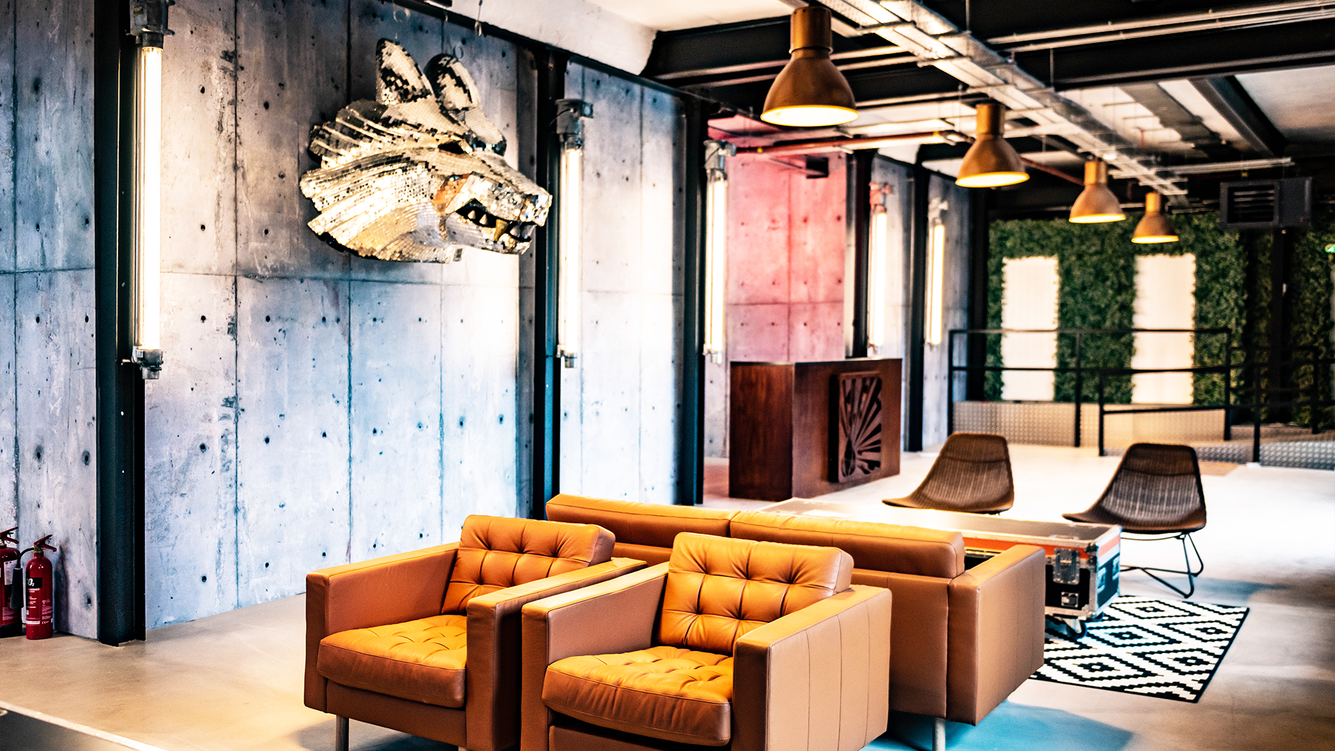 Modern loft-style interior with orange tufted sofas, patterned black and white rug, black chairs, concrete walls, gold hanging lights, and greenery wall section.