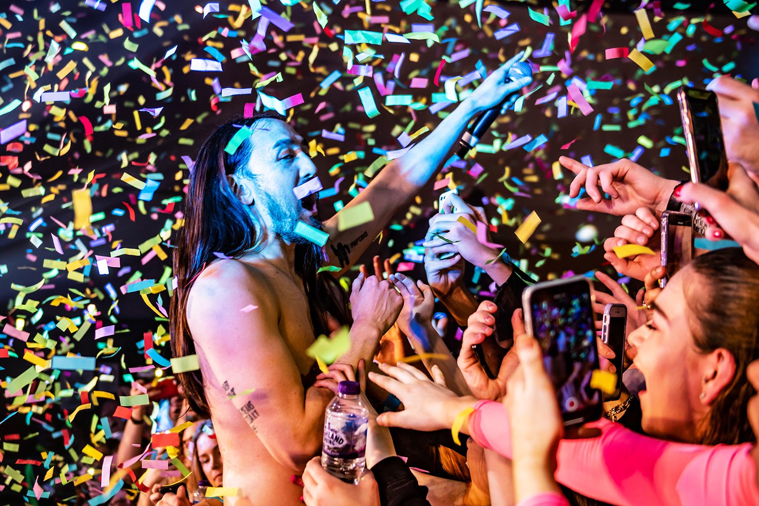 Performer celebrating at a concert or party with confetti falling around, surrounded by fans taking photos and reaching out.