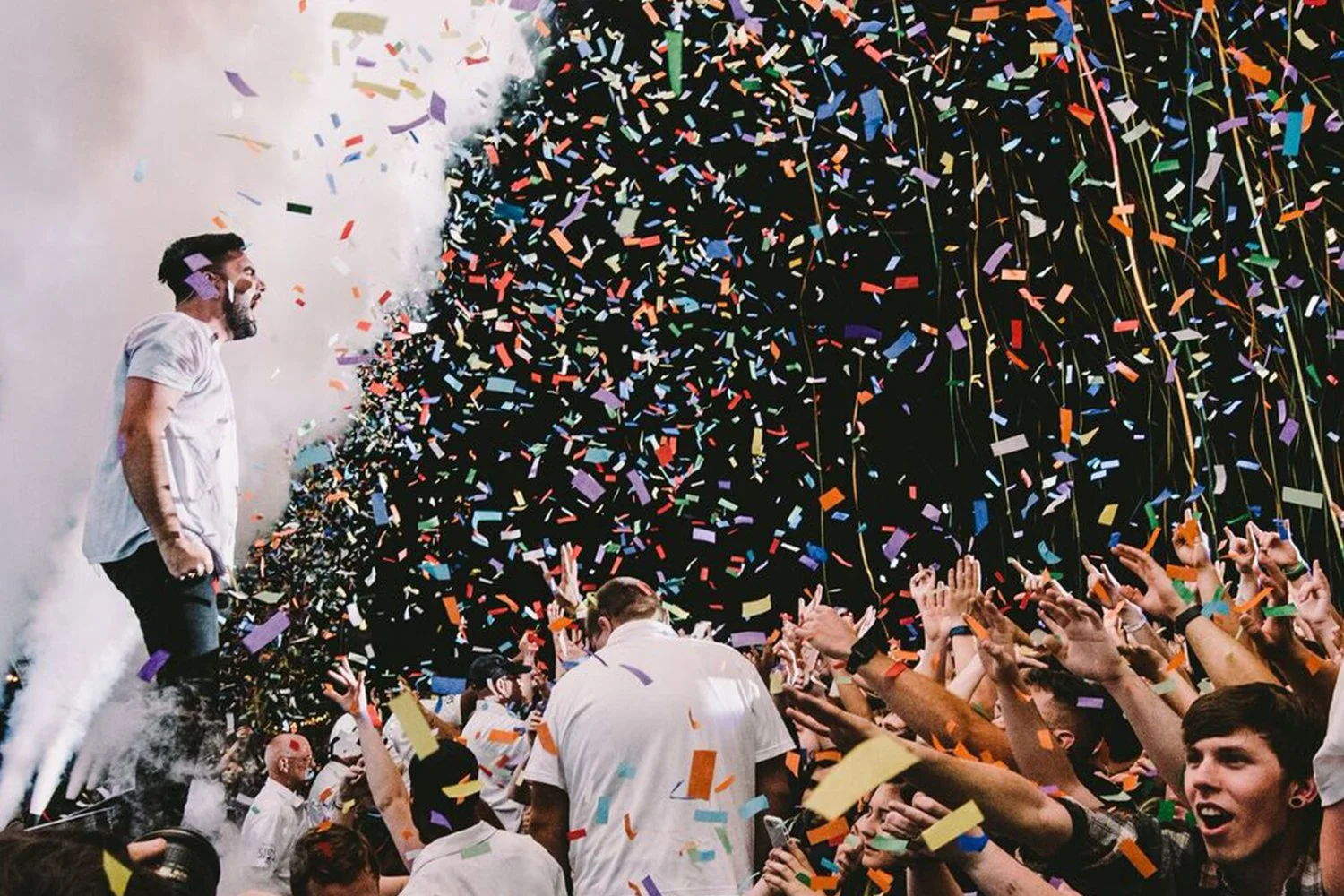 A man crowd surfing at a party with confetti falling around him and people reaching out to touch him, celebrating.