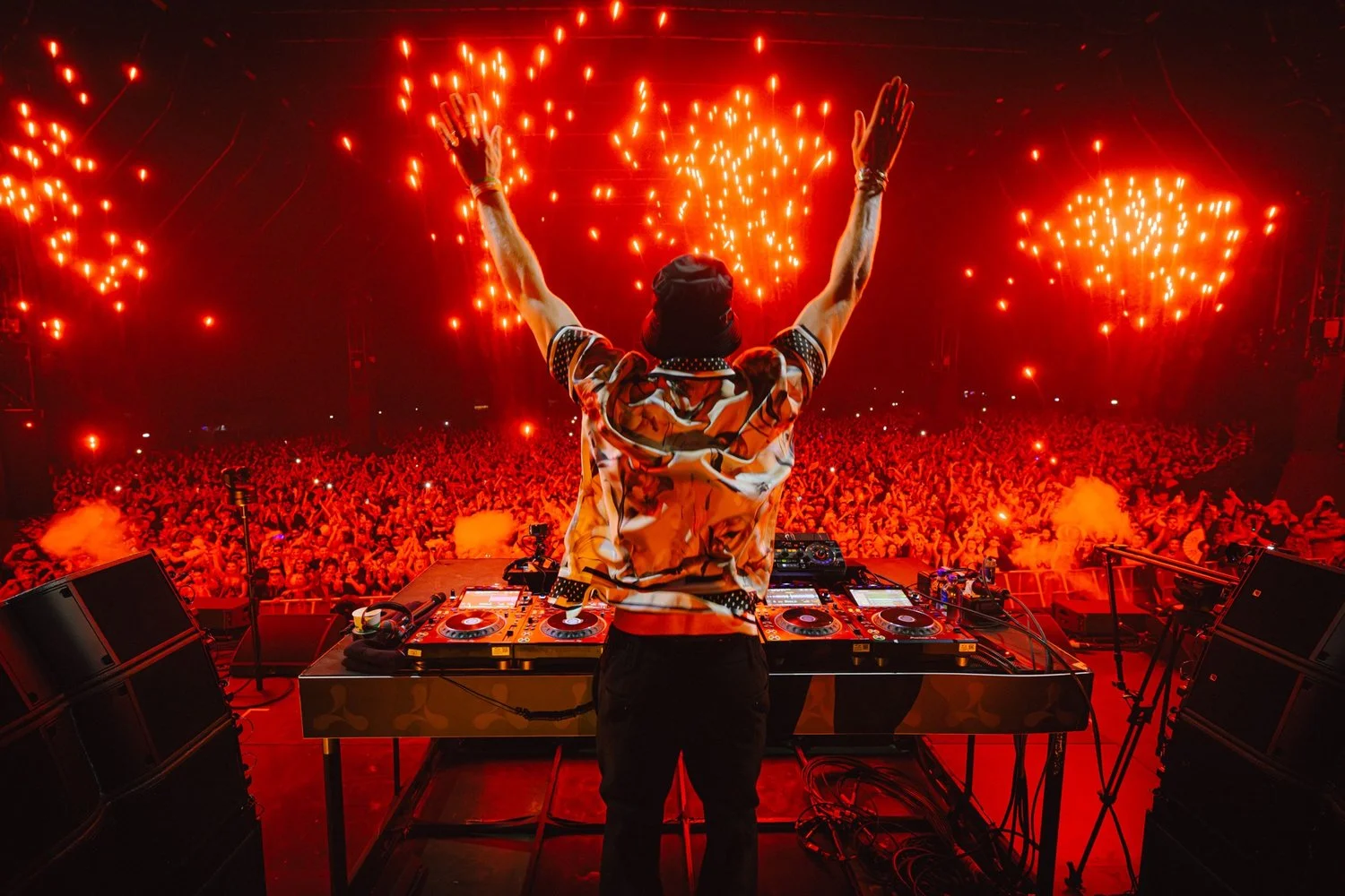DJ performing at a concert with a large crowd and red lighting, with fireworks in the background.