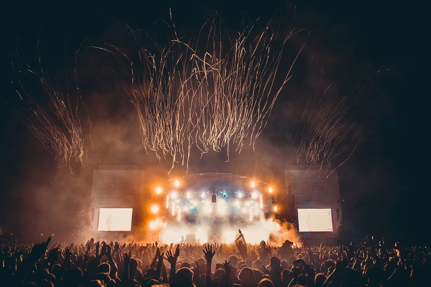 A large crowd at a concert or festival at night with fireworks in the sky, stage lights, and people raising their hands.