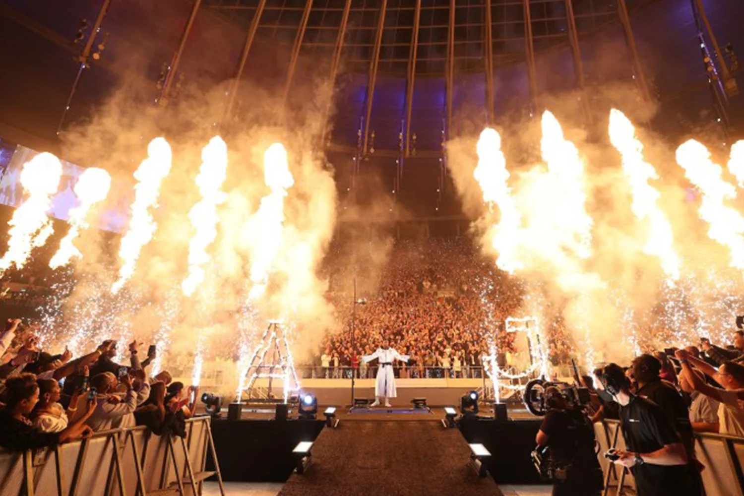 Performance at an indoor event with pyrotechnics and fireworks surrounding a person on stage, with a large audience and crew members capturing the event.