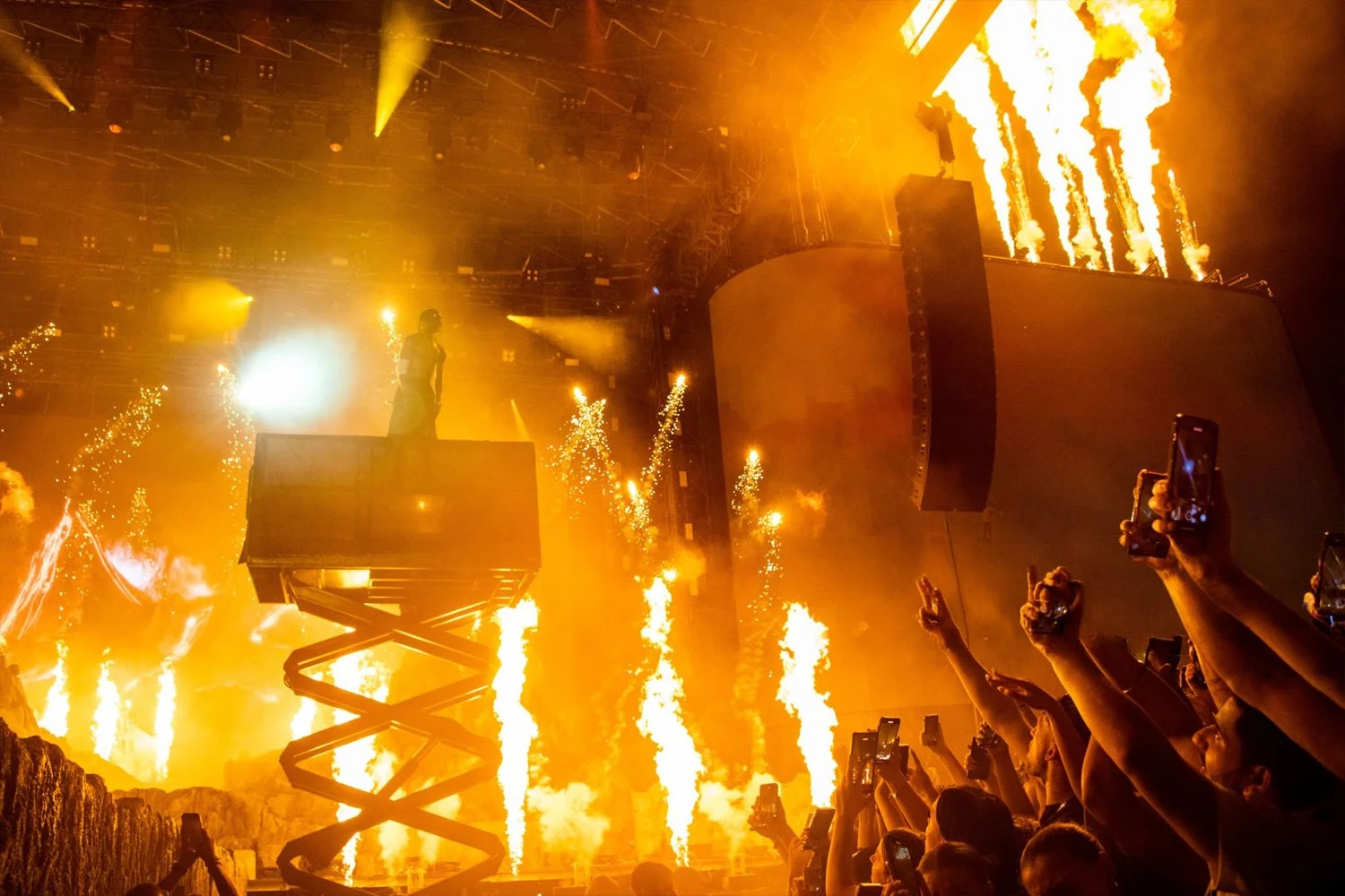 A concert scene with pyro and bright lights, a performer on a raised platform, and an audience holding up phones to record the event.