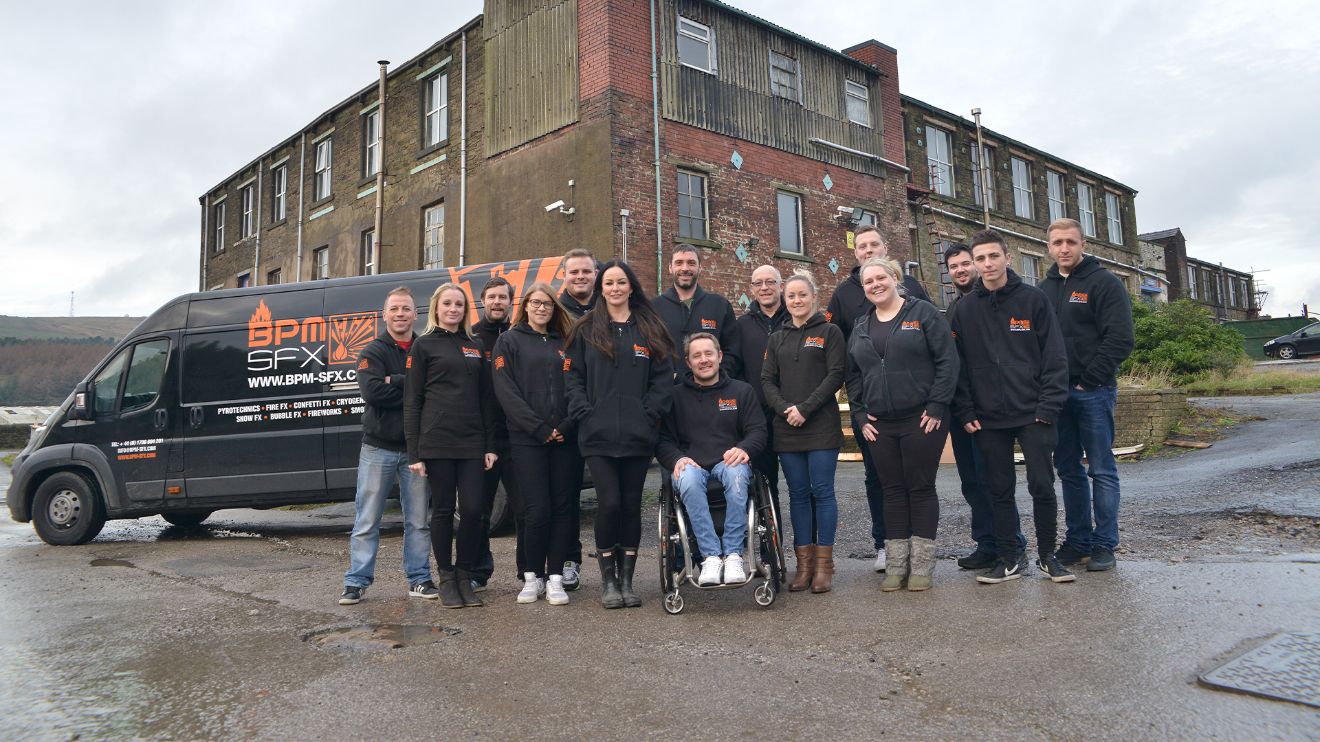 Group of people standing outdoors in front of a black van with business logos, near an old brick building, on a cloudy day.