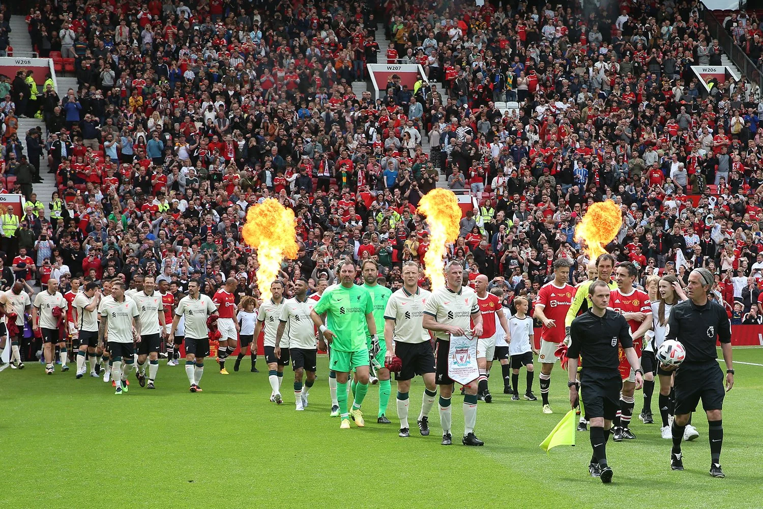 Soccer players and referees walking onto the field before a match, with flames shooting up in the background, and audience sitting in stadium seats