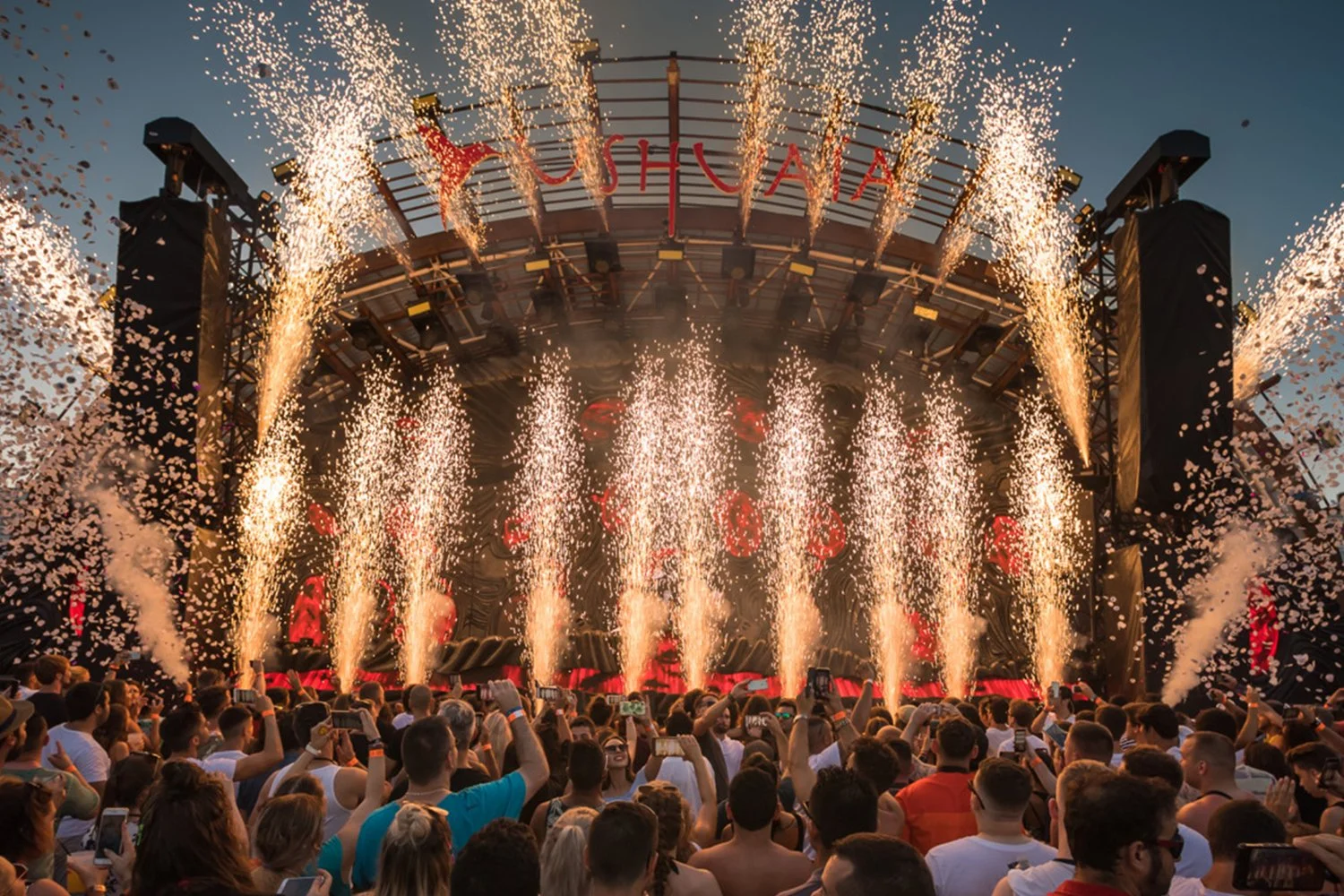 A live outdoor concert with fireworks and confetti, a large crowd watching and taking pictures, a stage with a curved roof, and the word 'YUSHUAIA' displayed at the top.