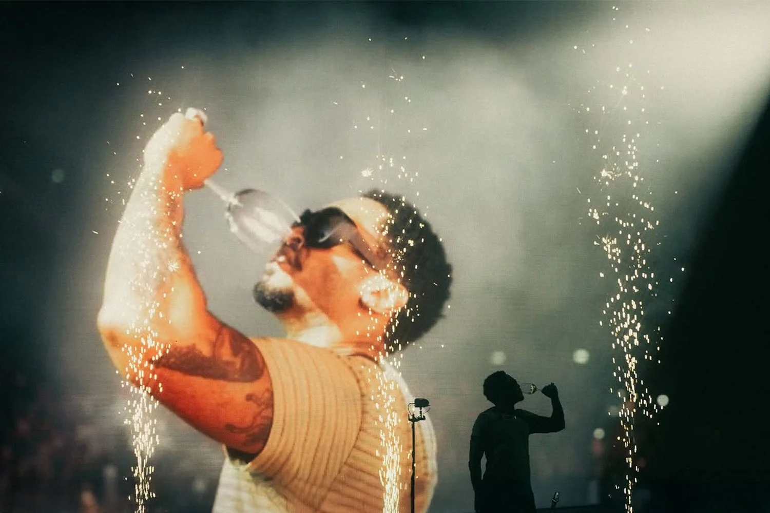 A man with tattoos on his arm and wearing sunglasses is drinking from a bottle amid fireworks and sparks, with a silhouette of another person in the background.