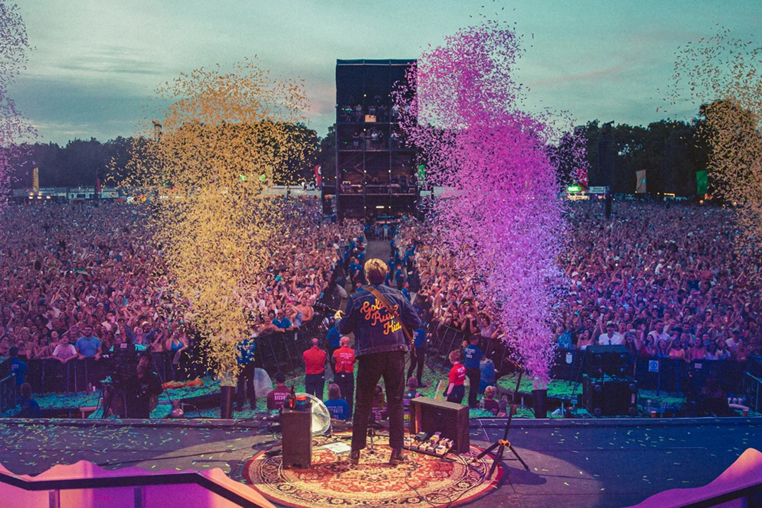A musician performs on stage at a large outdoor concert during sunset, with a massive crowd of thousands of people celebrating, colored confetti in the air, and a backdrop of trees and a darkening sky.