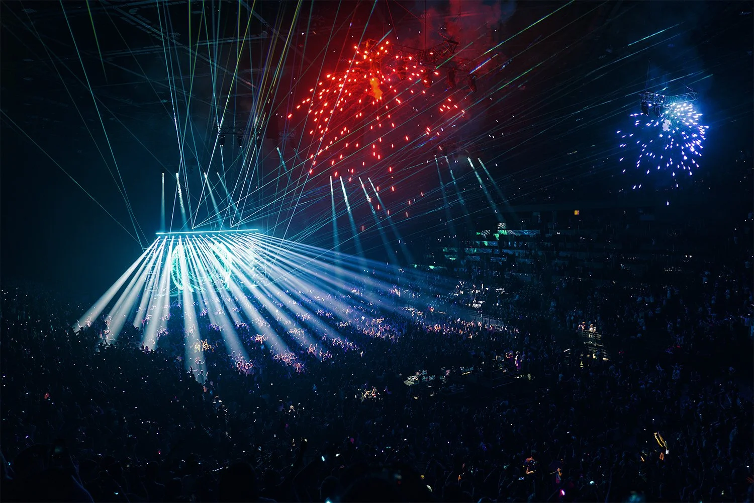 A concert with bright stage lights, laser beams, and colorful fireworks illuminating a large crowd.