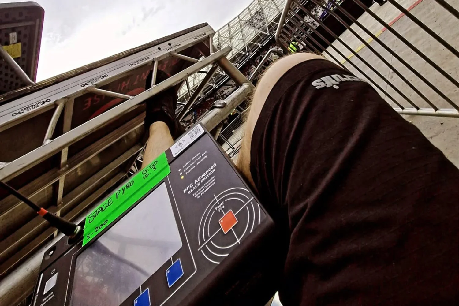 Photograph taken from a low angle showing a person wearing black shorts at a shooting range. Someone is aiming a pistol at a target downrange, with digital display screens and safety barriers visible.
