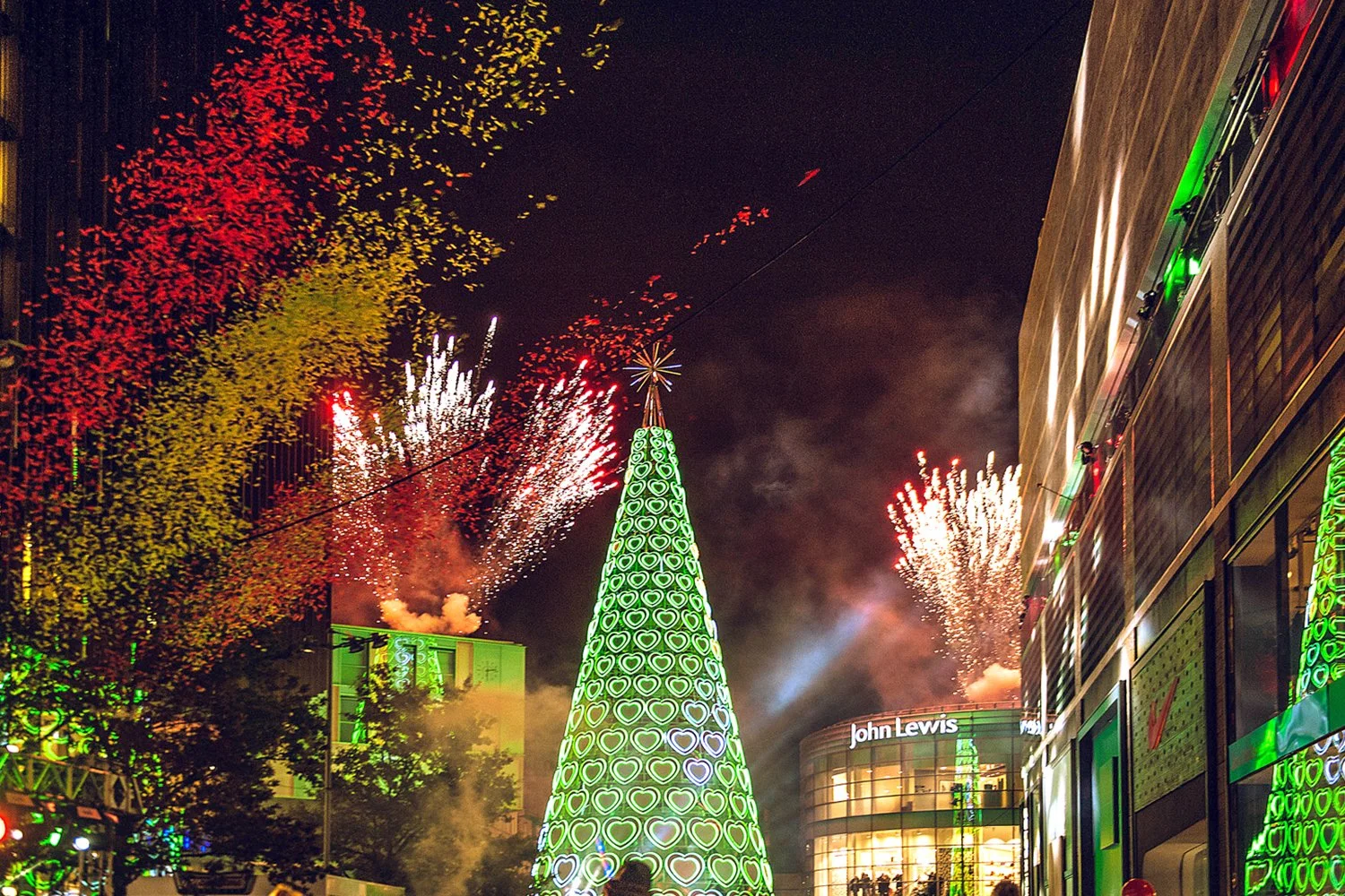 Nighttime view of a city street decorated for the holidays with a large illuminated Christmas tree topped with a star and fireworks exploding in the sky. Building facades are lit with green and other colorful lights, and a storefront with the sign 'John Lewis' is visible.