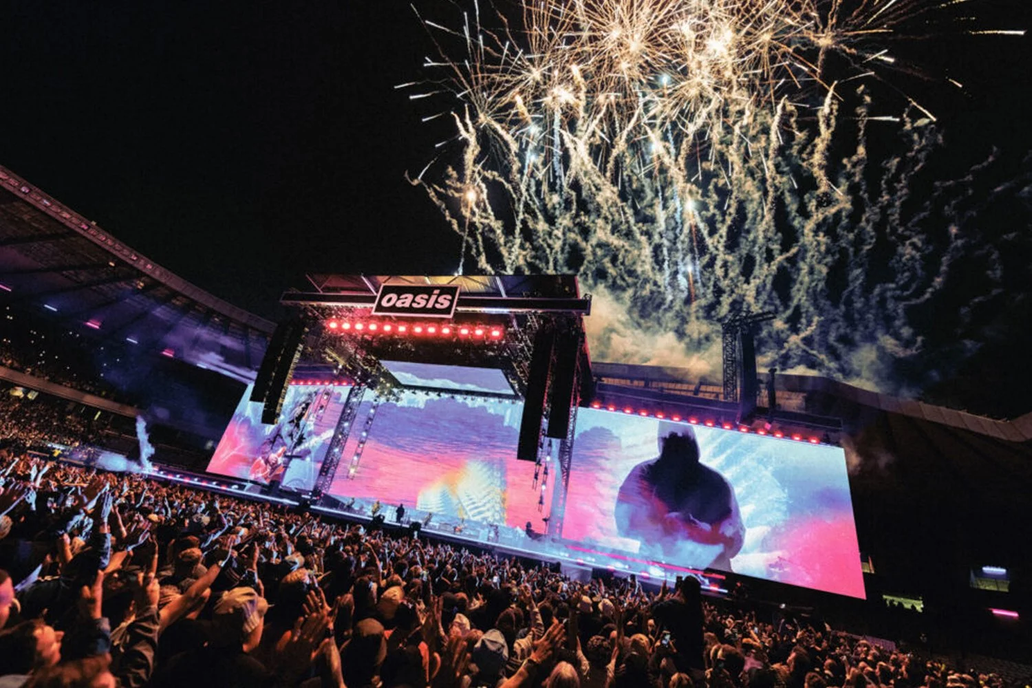 Crowd attending a concert at Oasis stadium during fireworks display at night.