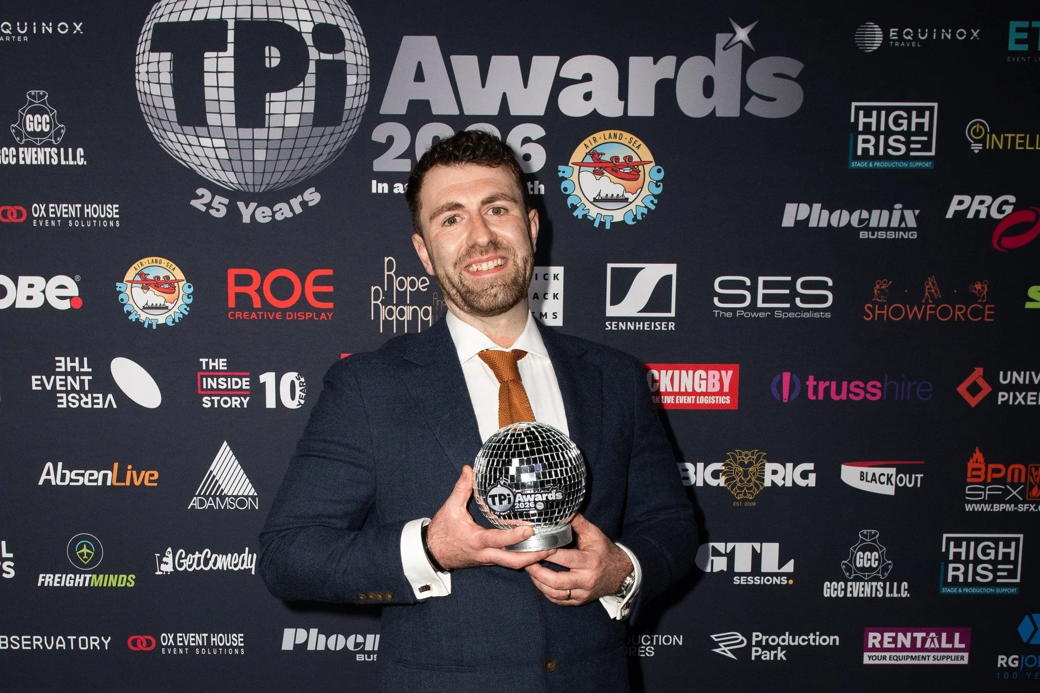 A man in a suit holding a disco ball-shaped award at an awards event. The backdrop features various sponsor logos and text indicating the event name, 'TIPA Awards 2026,' and celebrating 25 years.