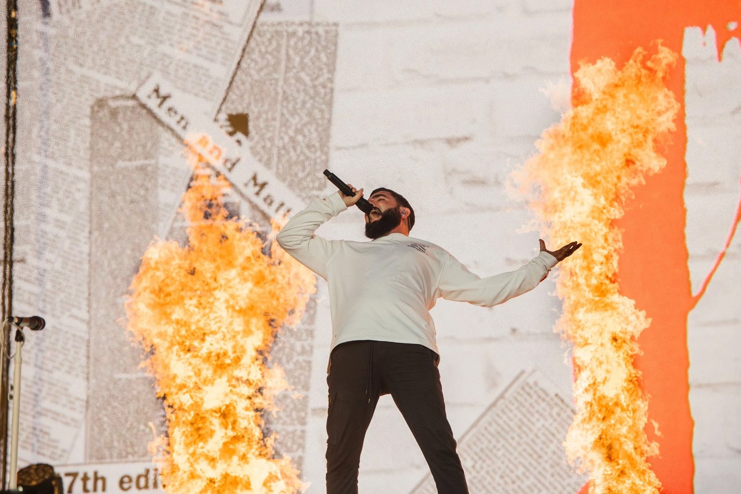 A man with a beard wearing a light-colored jacket singing into a microphone on stage, with flames on either side and a background featuring a large collage of newspaper articles.