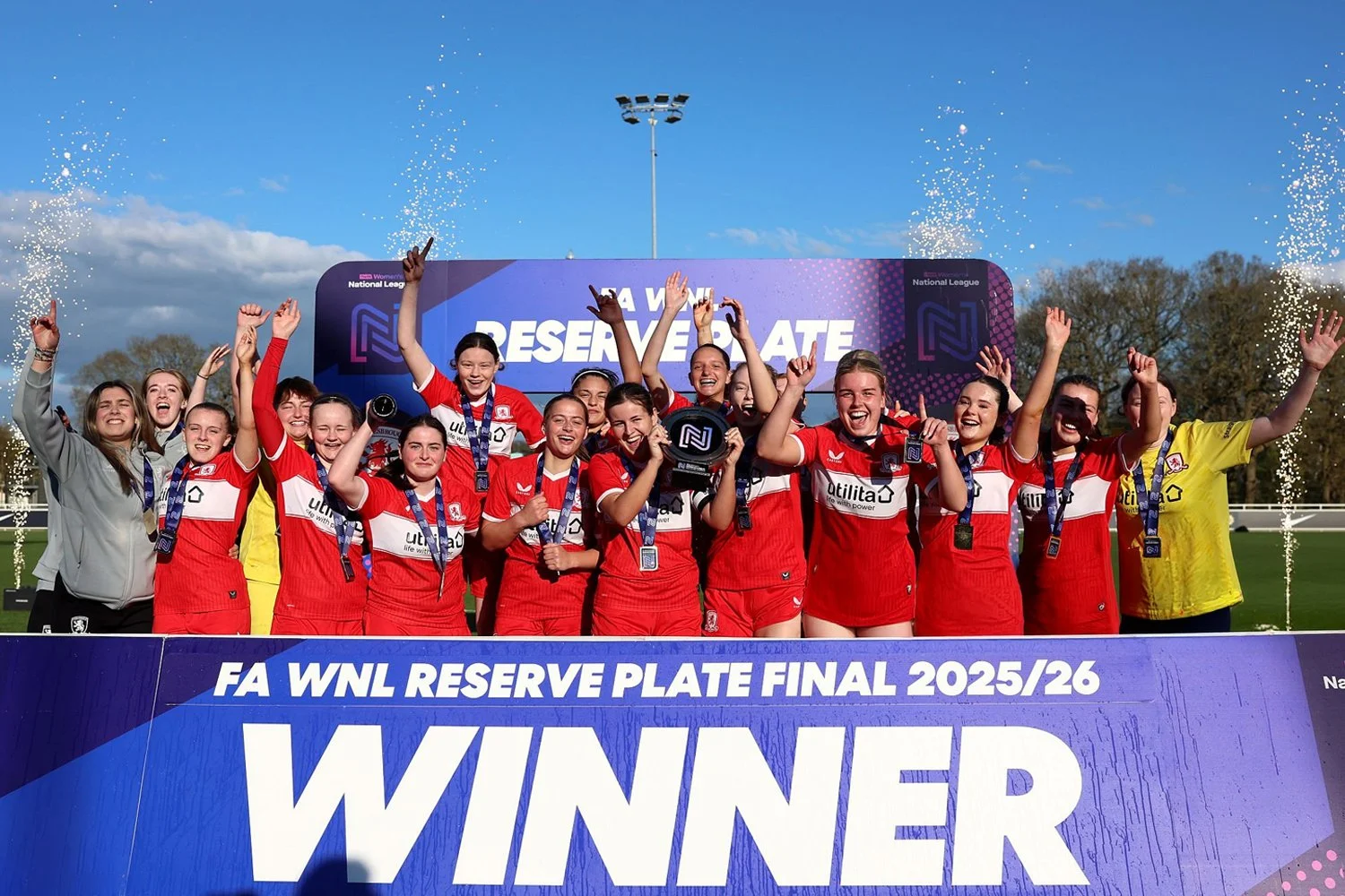 A team of female soccer players celebrating their victory at the FA WNL Reserve Plate Final 2025/26, holding a trophy with medals around their necks, on a podium with a large banner that reads 'WINNER' and 'FA WNL RESERVE PLATE FINAL 2025/26'.