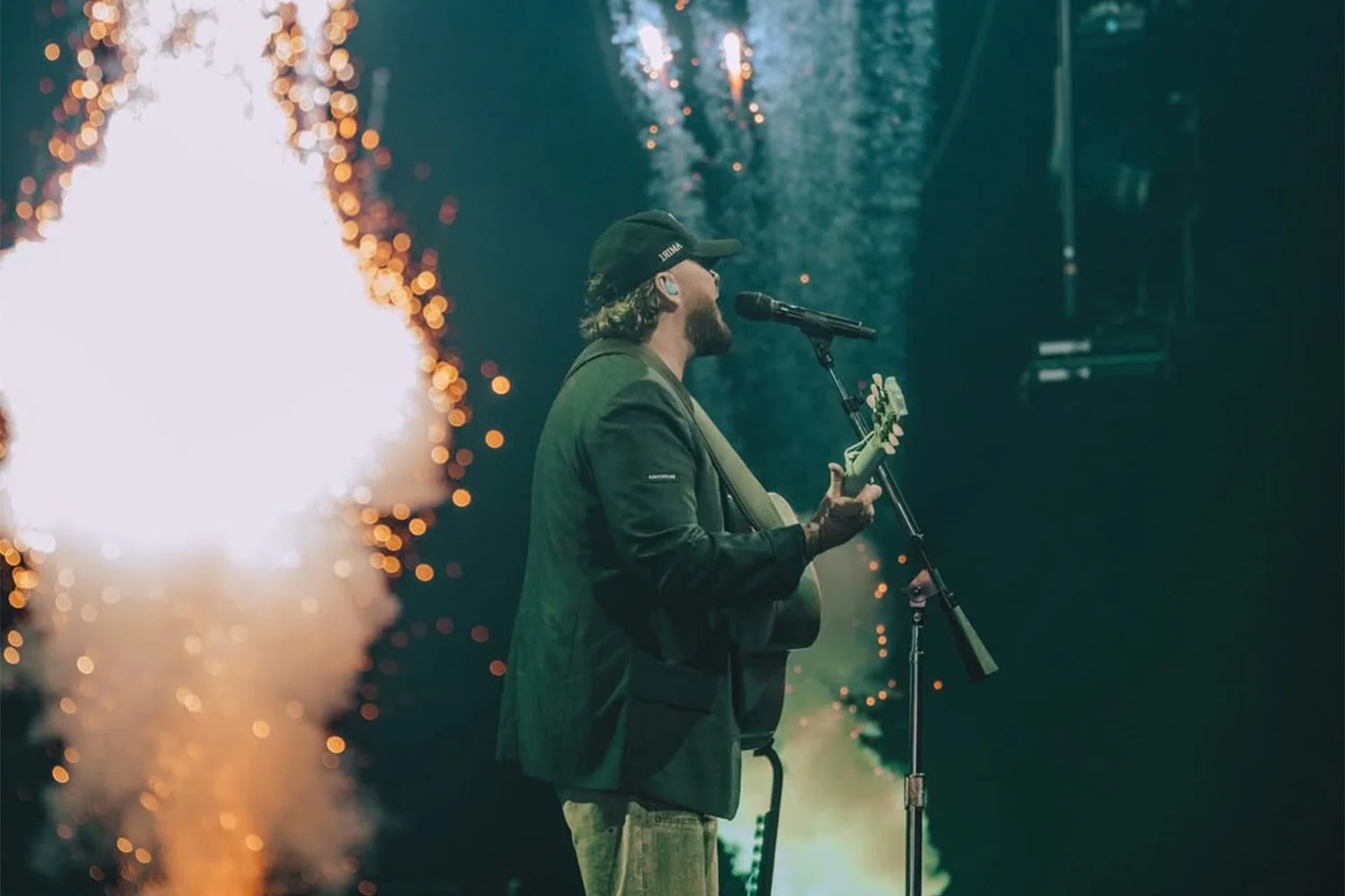 A man wearing a black cap and jacket, singing into a microphone while playing an acoustic guitar on stage, surrounded by fireworks.