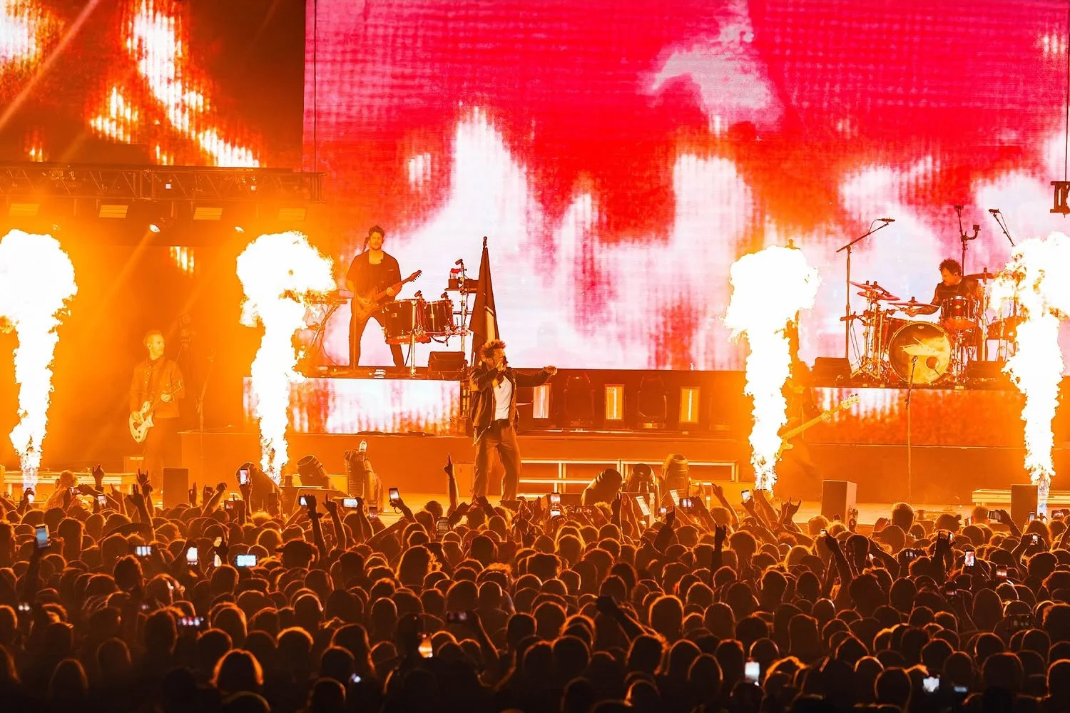 Live concert with a large audience, band performing on stage with pyrotechnics and a digital screen displaying red and white abstract visuals.