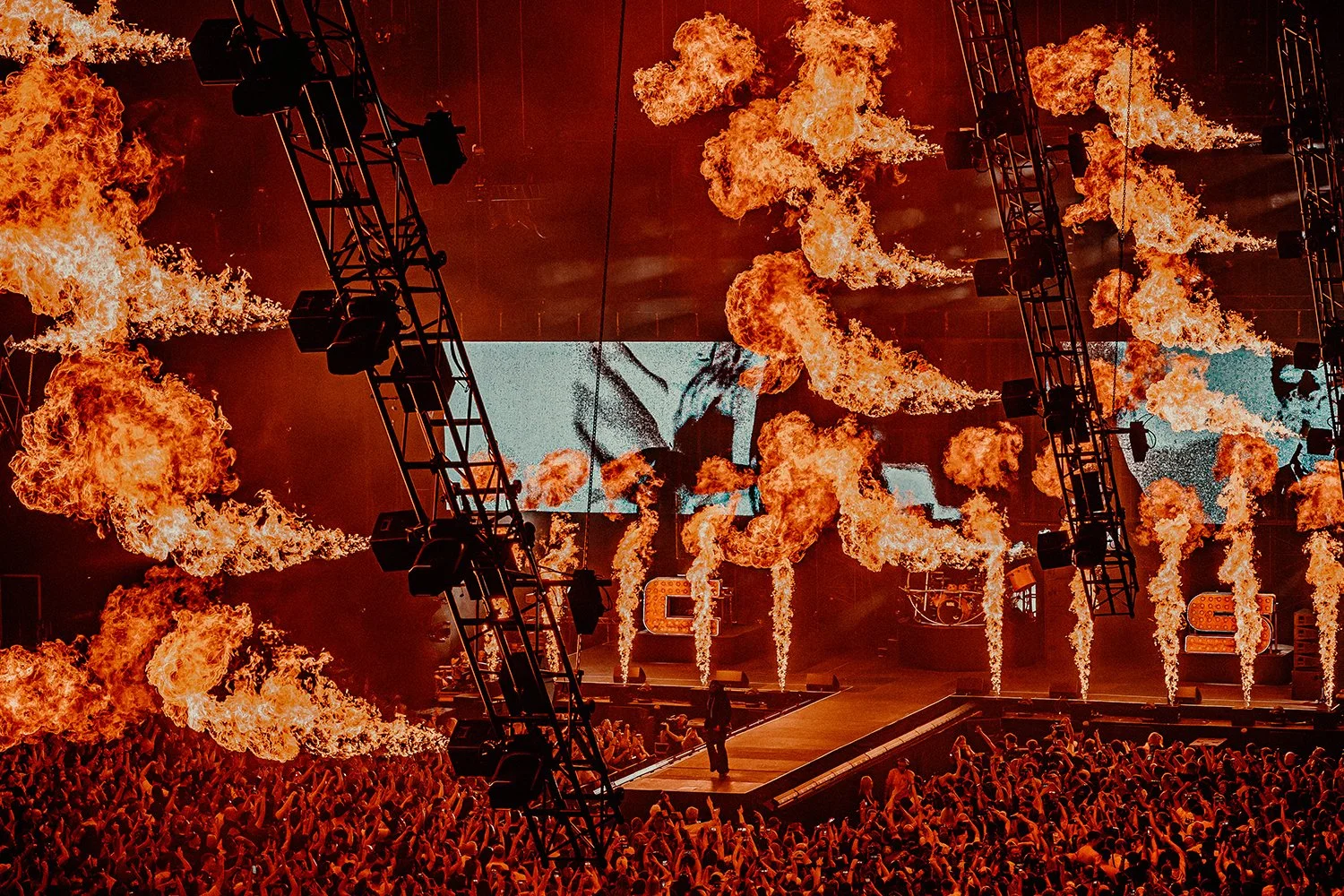 A large concert stage with multiple fire effects and flames shooting upward, a big crowd in front, and a large screen displaying visuals in the background.