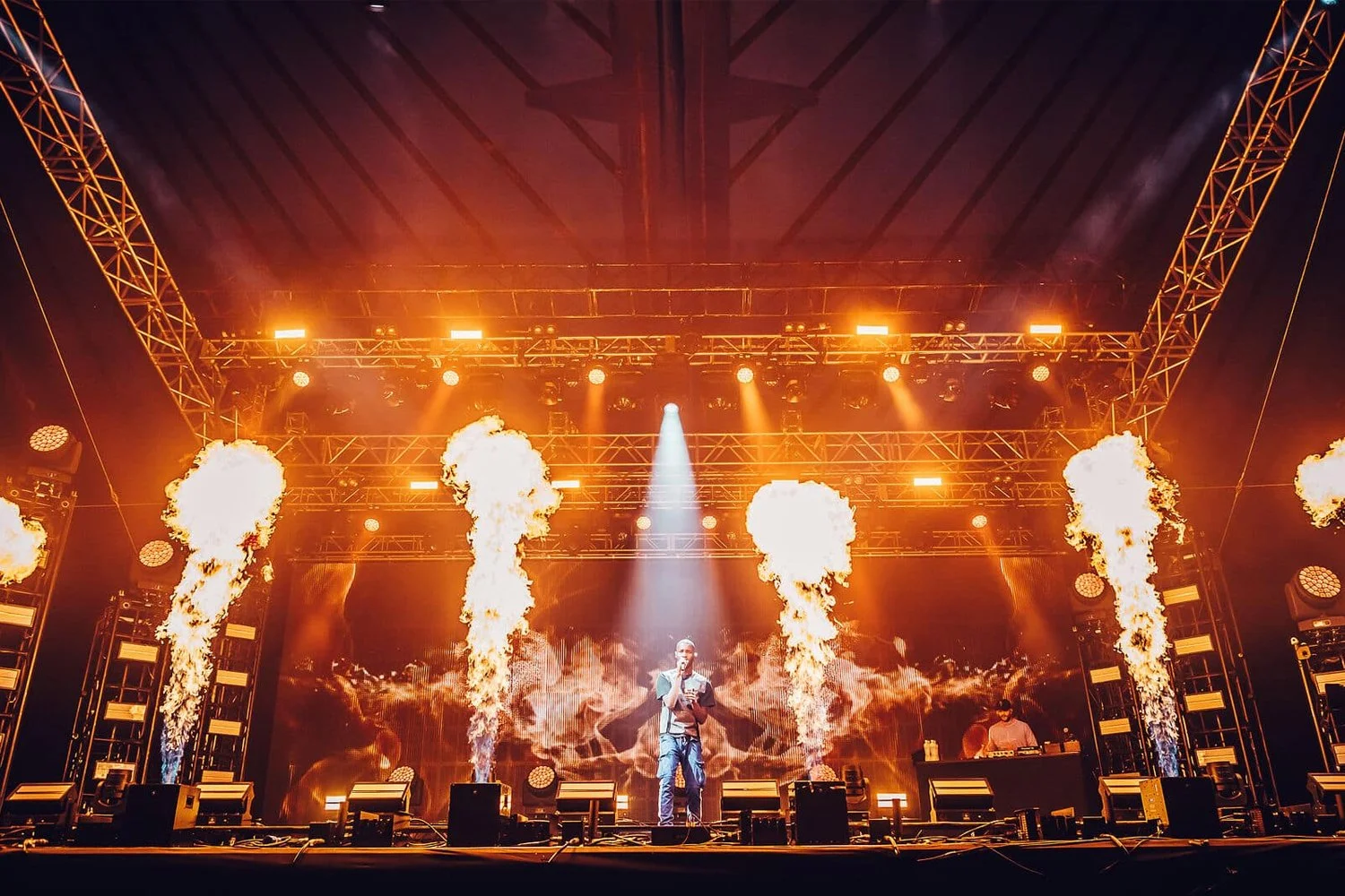 A male performer on stage at a concert with fire effects and bright orange lights, with a DJ in the background.