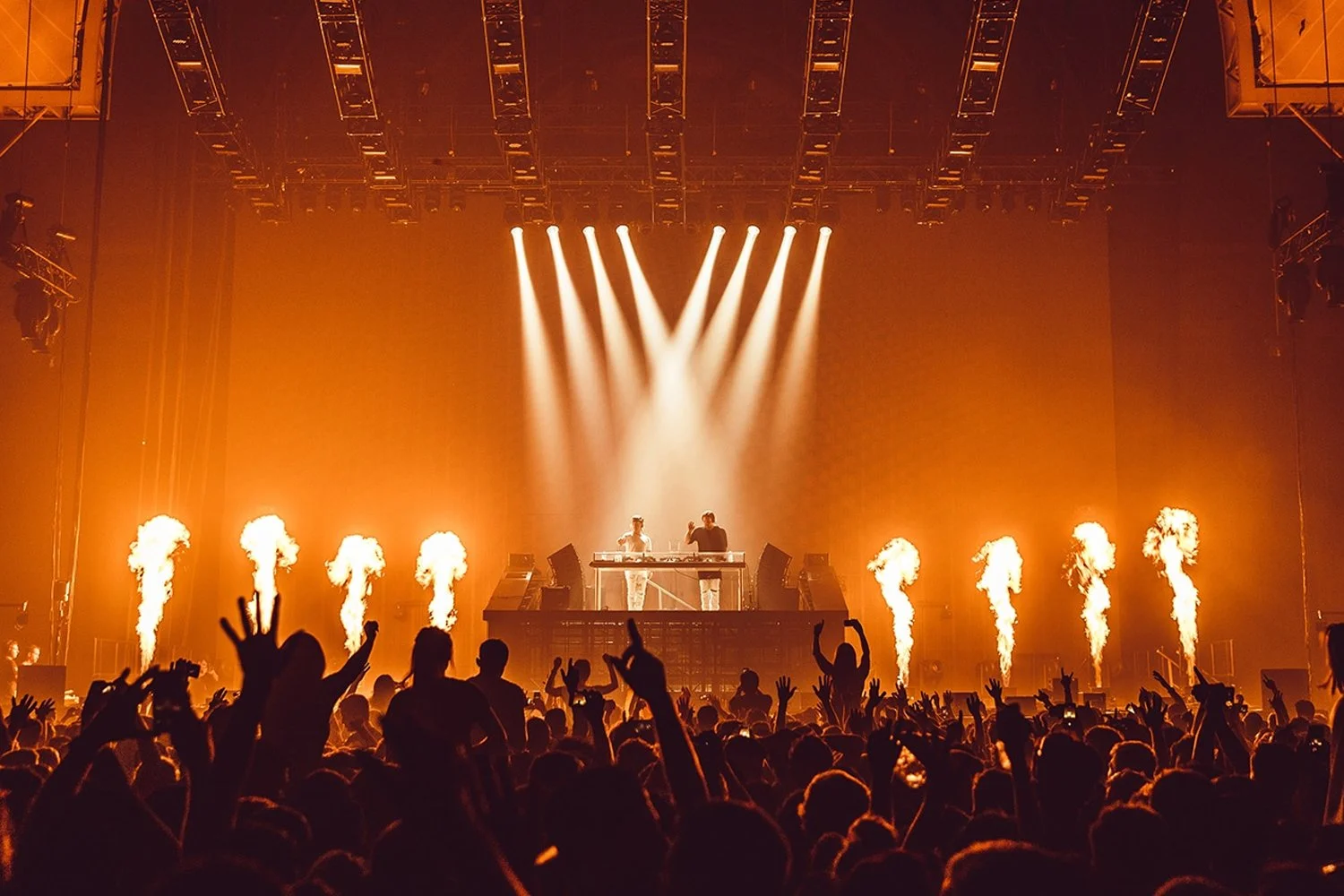 A concert scene with a DJ on stage, surrounded by flames and bright lights, with an audience dancing and raising their hands.