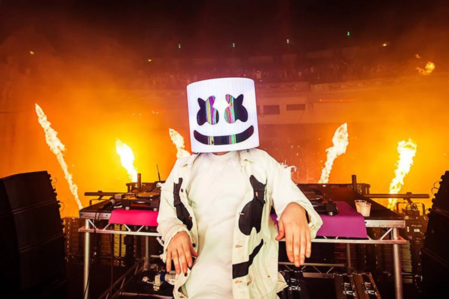 DJ Marshmello performing on stage with fire effects and pyrotechnics in the background.