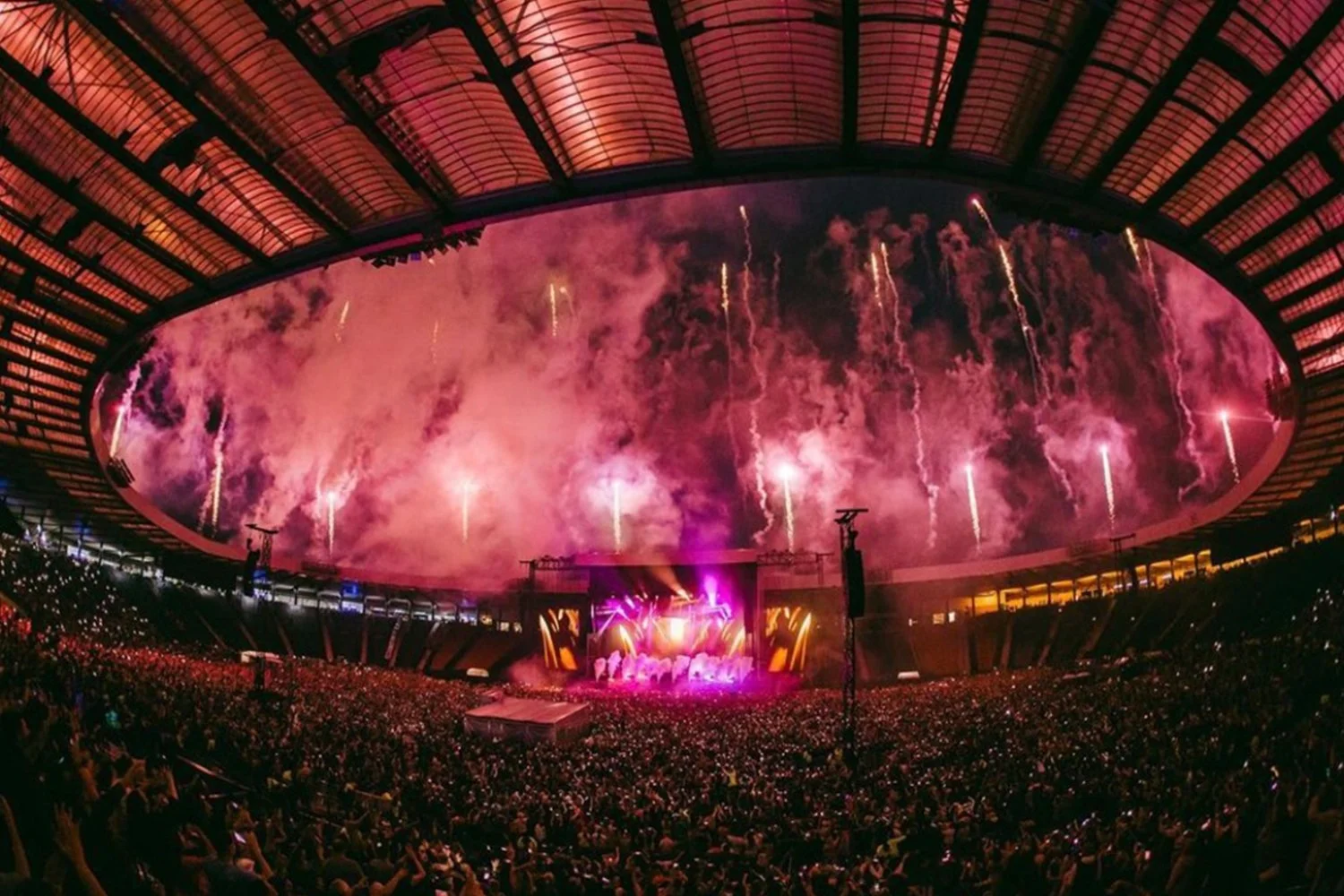 A large indoor stadium with a curved roof, filled with a packed crowd, and a stage with bright purple and yellow lighting, fireworks and smoke in the background, creating a vibrant concert atmosphere.